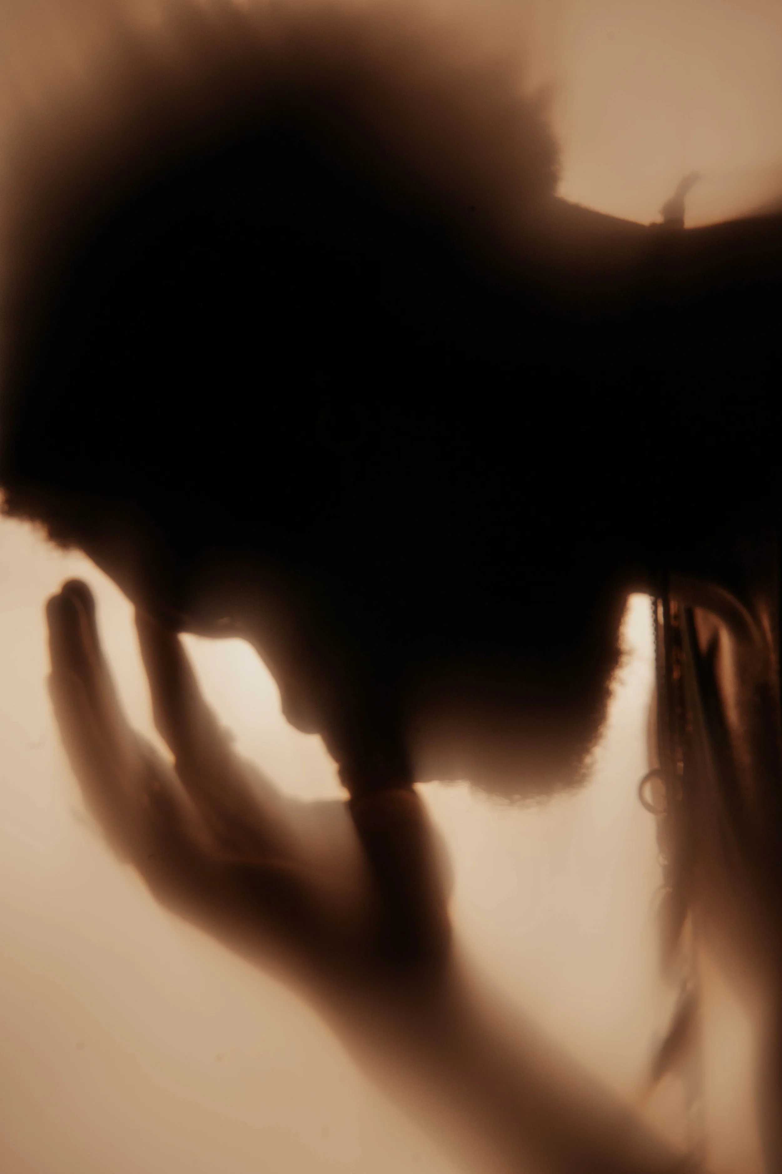 A close-up shot of a person's hand with their fingers extended, holding an object, with a blurry background that includes a silhouette of a person, possibly outdoors during sunset or sunrise.