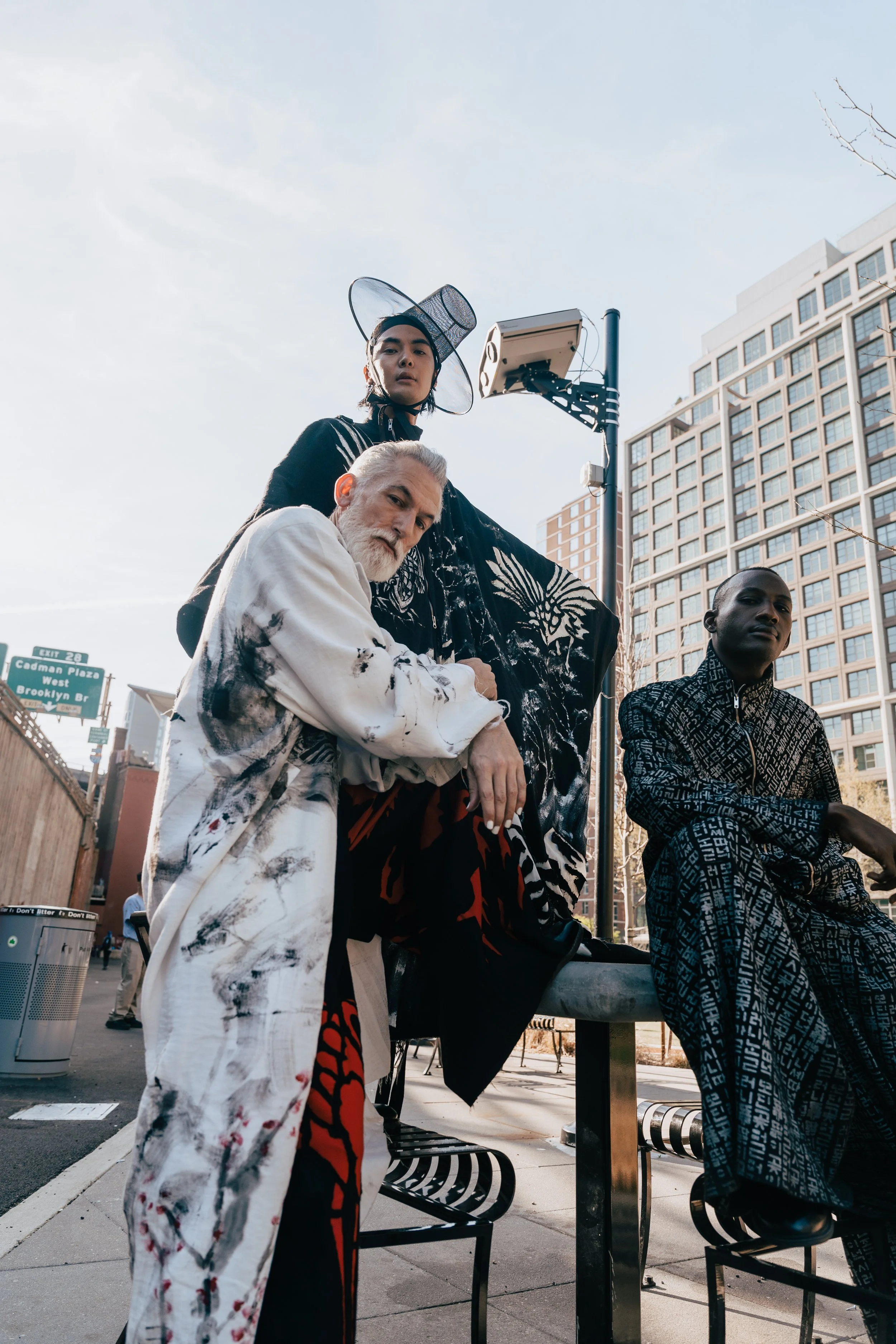 Three people are posing for a photo on a city street. The woman in the foreground wears a flowing white and black patterned coat and has gray hair. Behind her, a woman with dark hair is dressed in black with a wide-brimmed mesh hat. To the right, a m