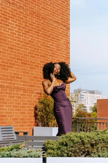 A woman in a purple strapless dress posing on a rooftop with a brick wall and cityscape background.