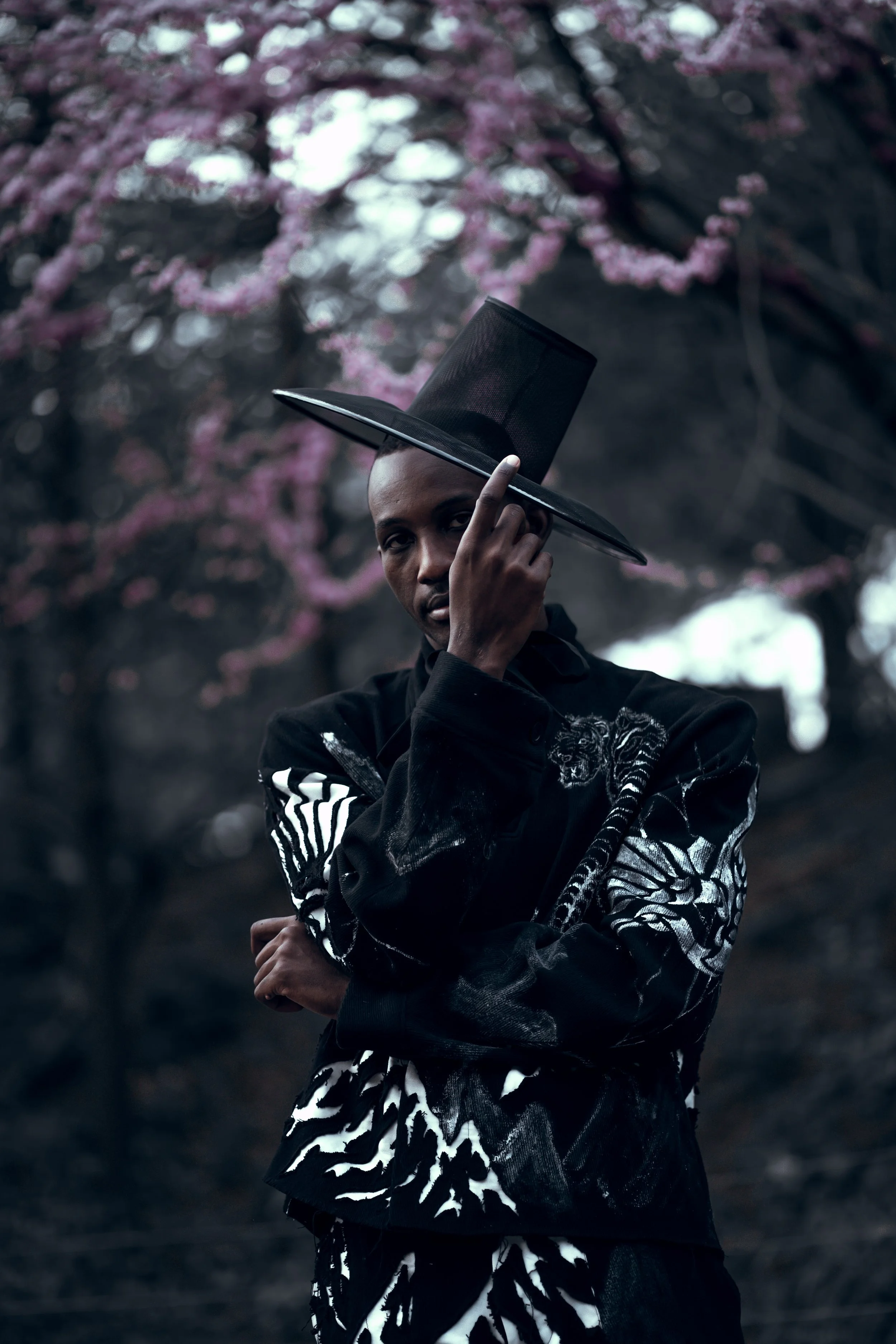 A person with dark skin is holding a large black hat with a wide brim, slightly tilted, while standing outdoors in front of a backdrop of pink flowering trees. The person is wearing a black jacket with artistic designs, including white tiger stripes and skull imagery, and has an intense expression.