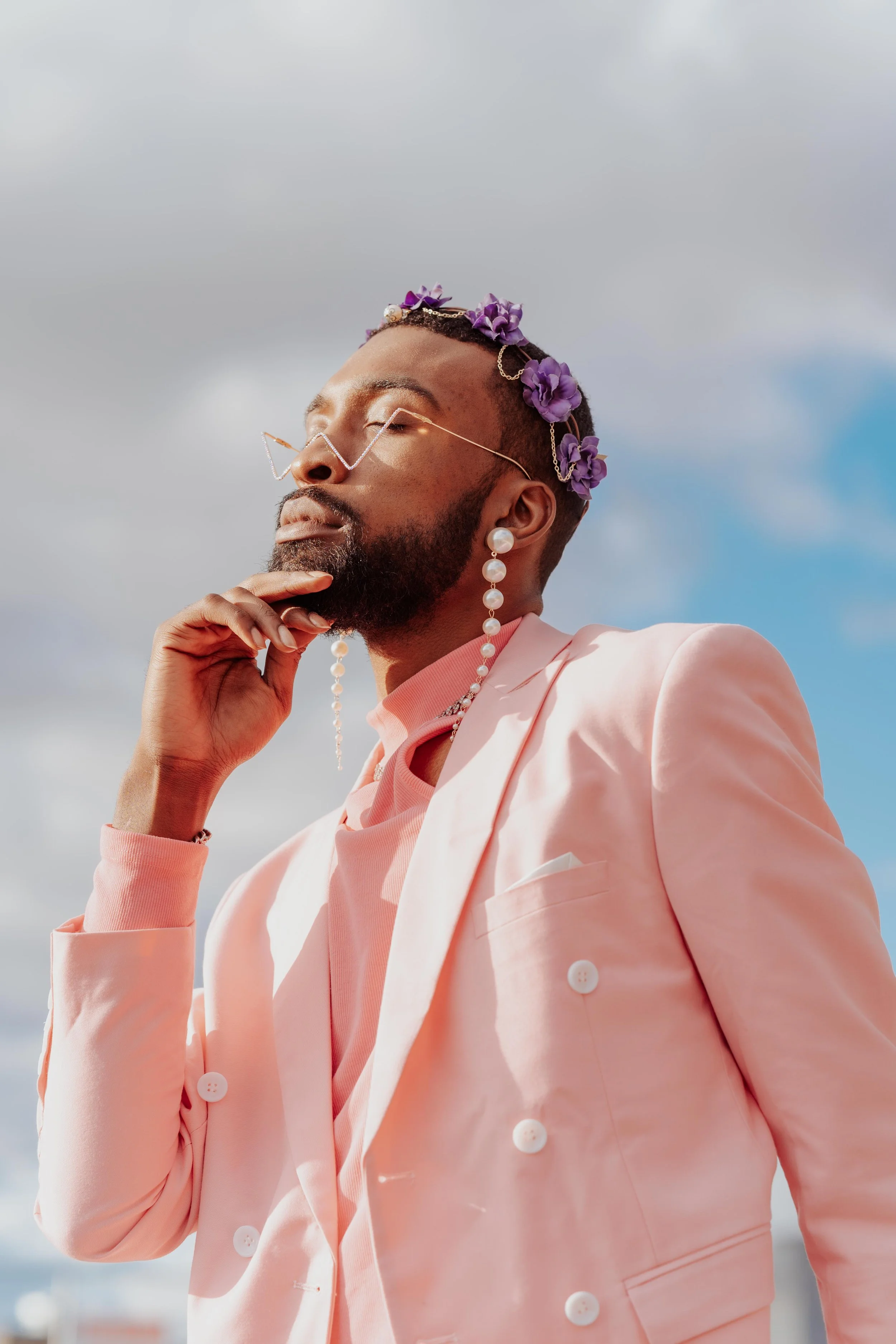 A person with a beard, closed eyes, and wearing glasses with a chain, adorned with purple flowers on their head, dressed in a pink blazer and shirt, posed outdoors against a cloudy sky.