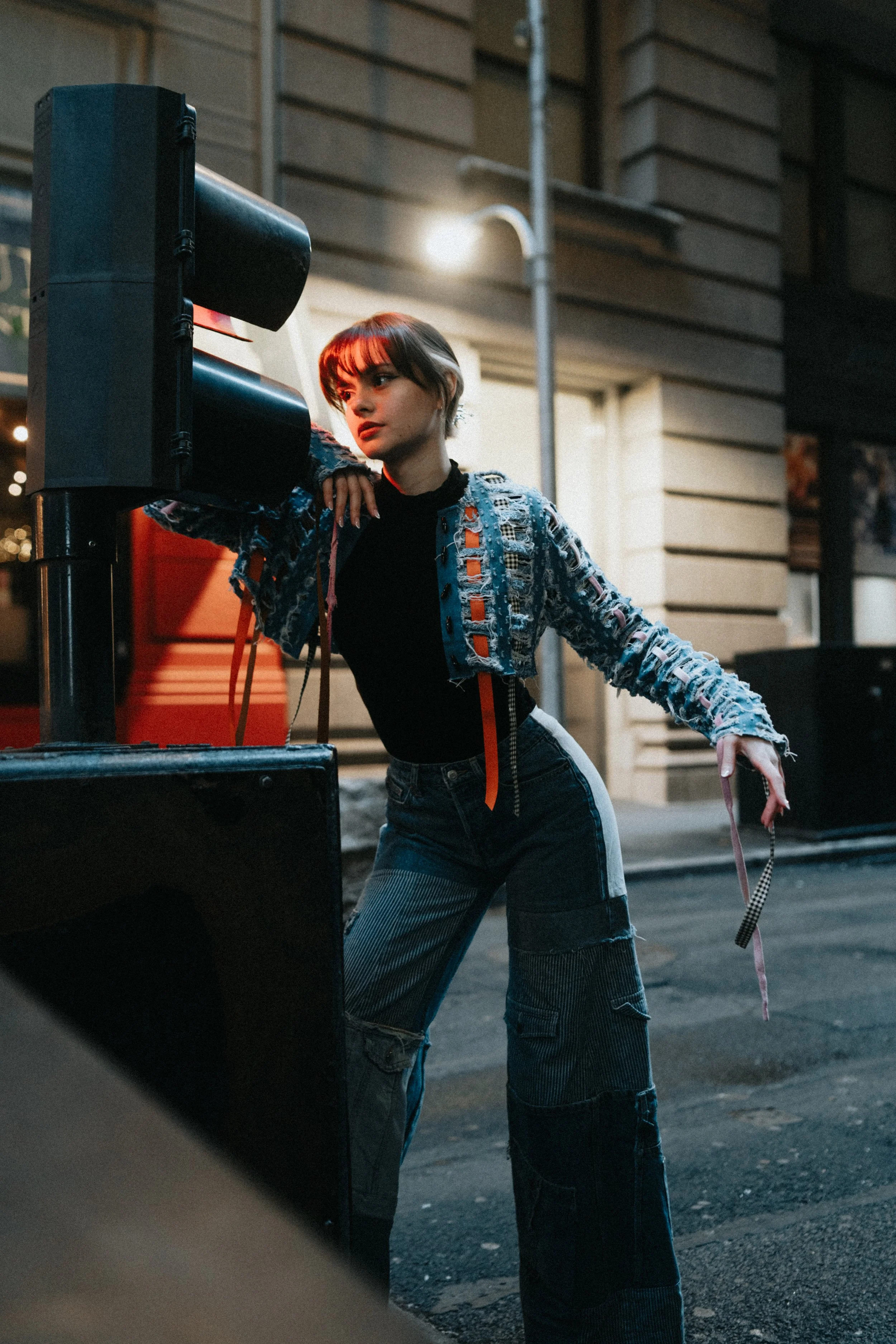 A young woman with short hair wearing a colorful, torn-up jacket and jeans, standing at a street corner at night, next to a traffic light, in an urban setting with buildings and streetlights.