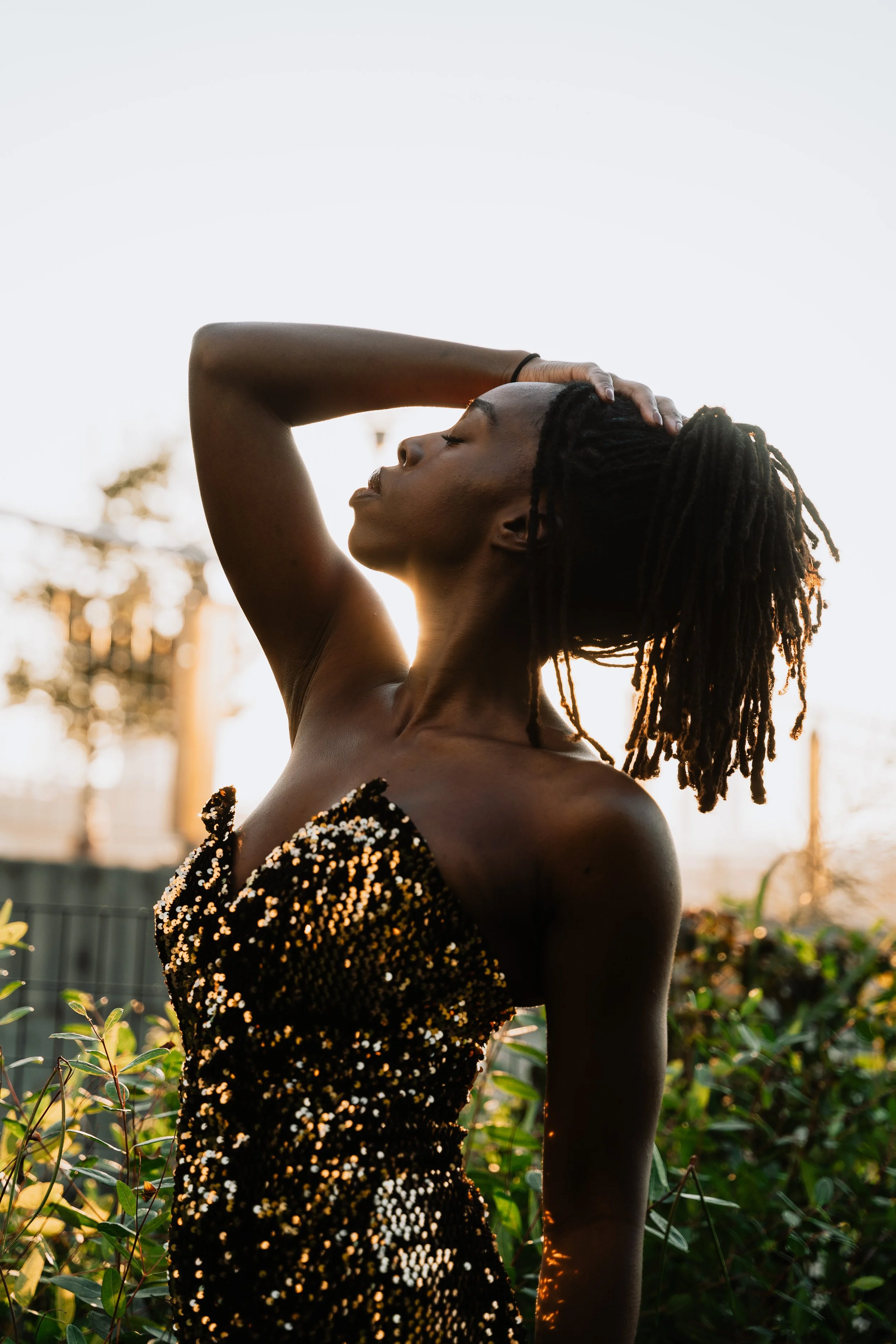 A woman with long dreadlocks and dark skin is standing outdoors with her eyes closed, one hand resting on her head, and wearing a black and gold sequined dress, backlit by the setting sun.