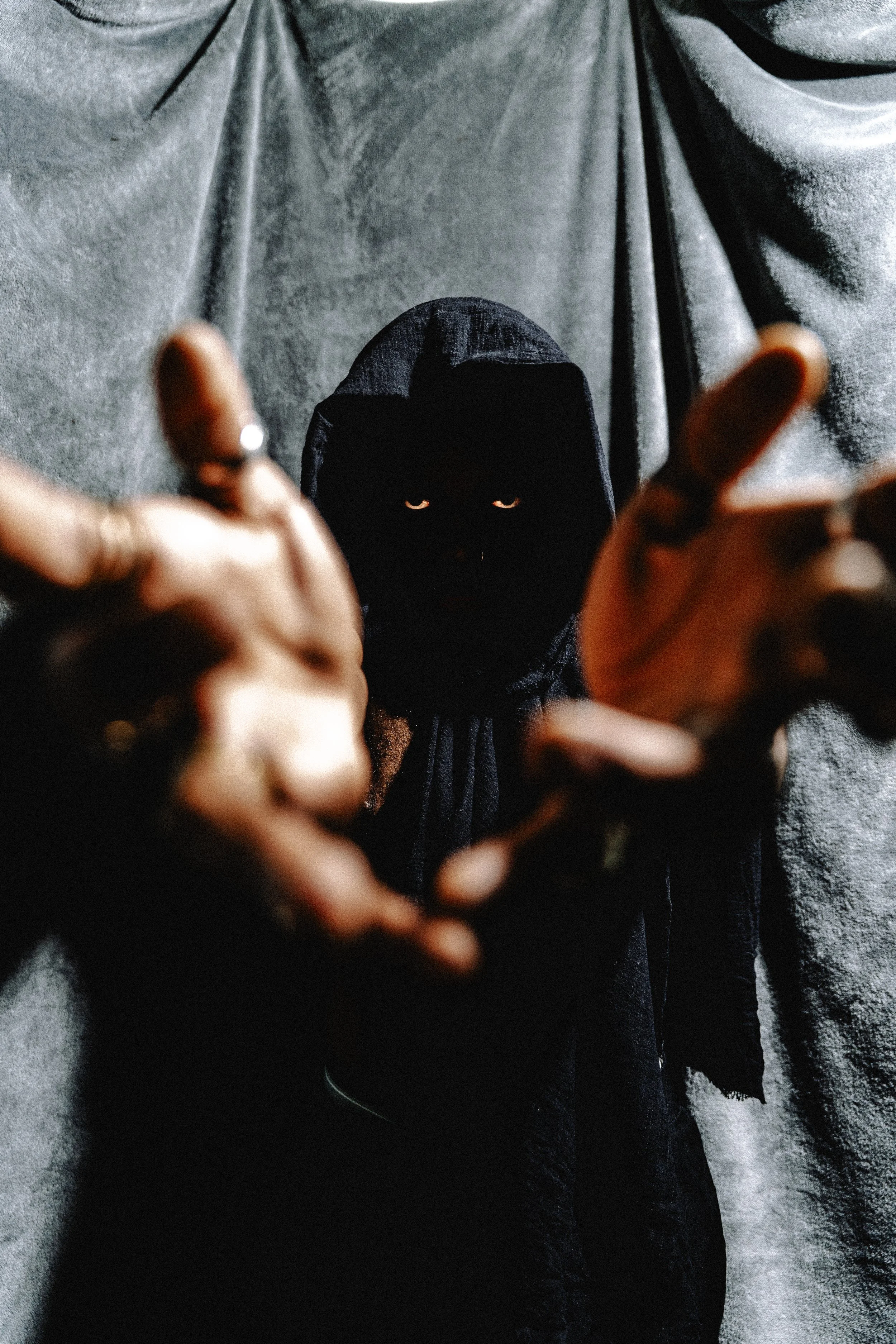 A person wearing a hoodie and making a hand gesture, with the face mostly obscured and eyes visible beneath the hood, standing against a textured wall.