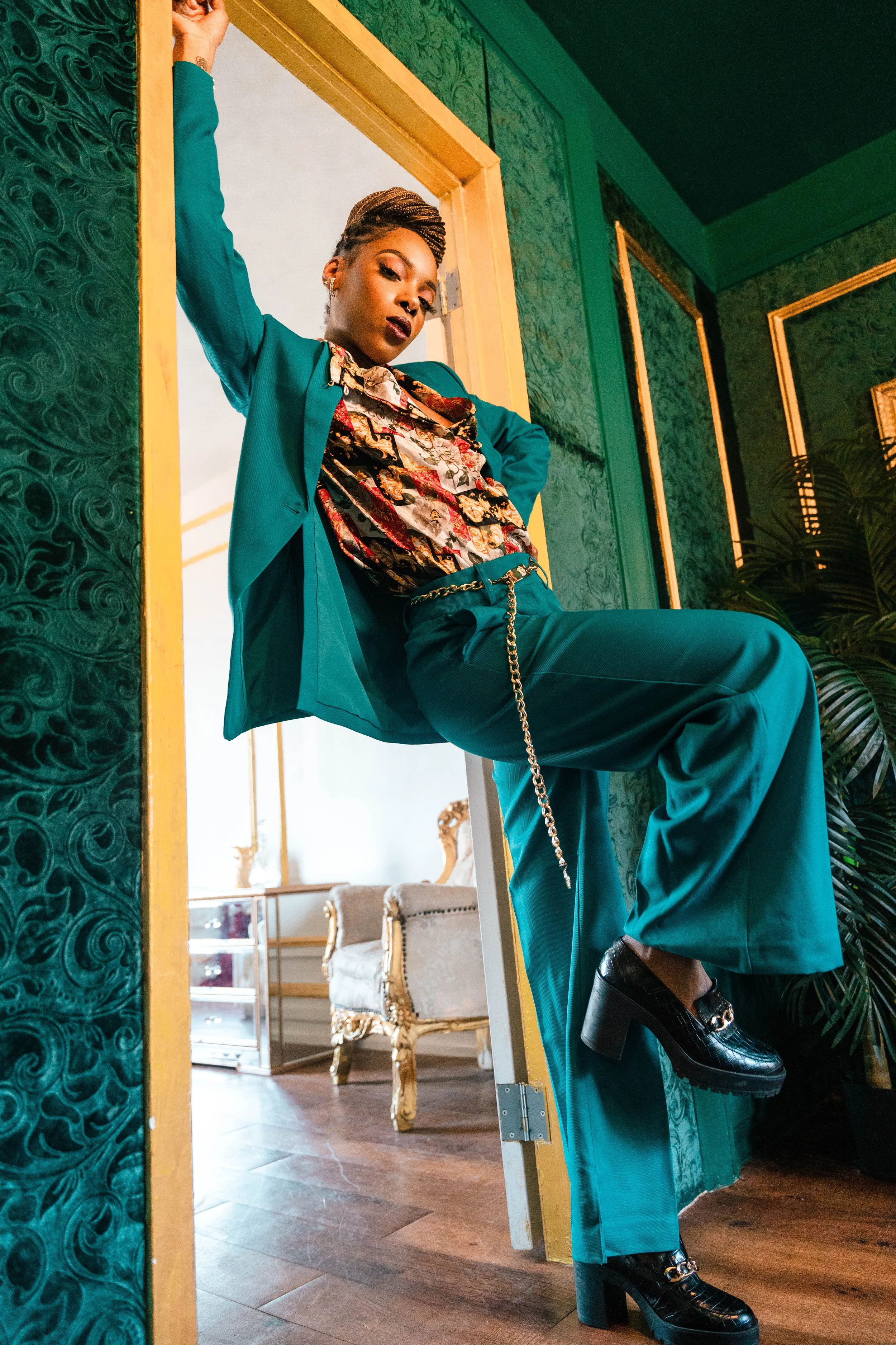 A woman in teal suit and patterned shirt poses with one arm on a doorway frame, displaying confidence, with a vintage styled room in the background.