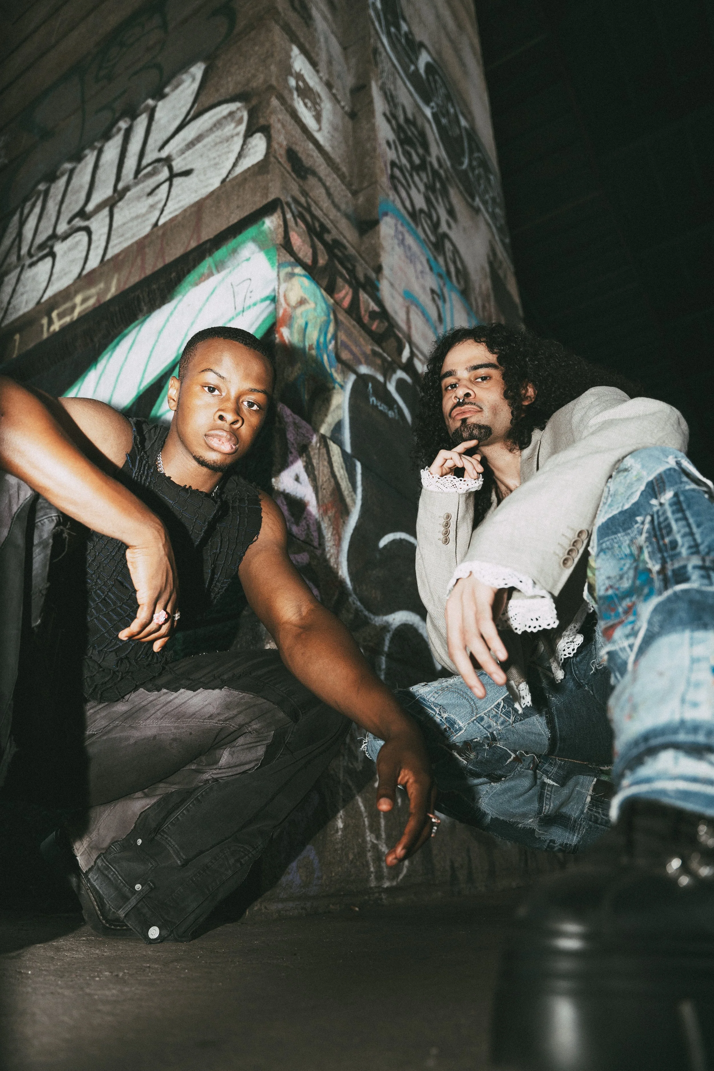 Two young men posing in an urban setting with graffiti-covered walls behind them. One has a shaved head and a serious expression, wearing a black sleeveless shirt. The other has curly hair, a nose piercing, and a contemplative look, wearing a light j