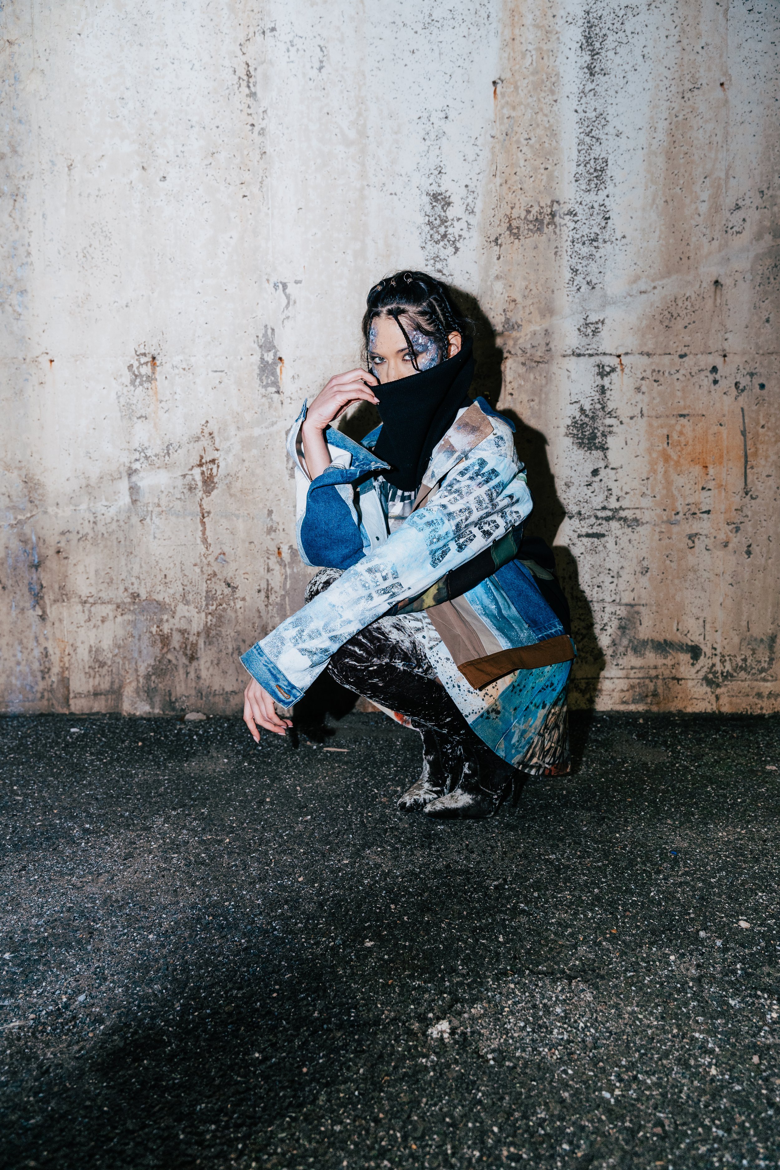 A person crouching on a wet asphalt street and posing against a weathered concrete wall, wearing a patchwork jacket, dark pants, patterned boots, and a black face covering.