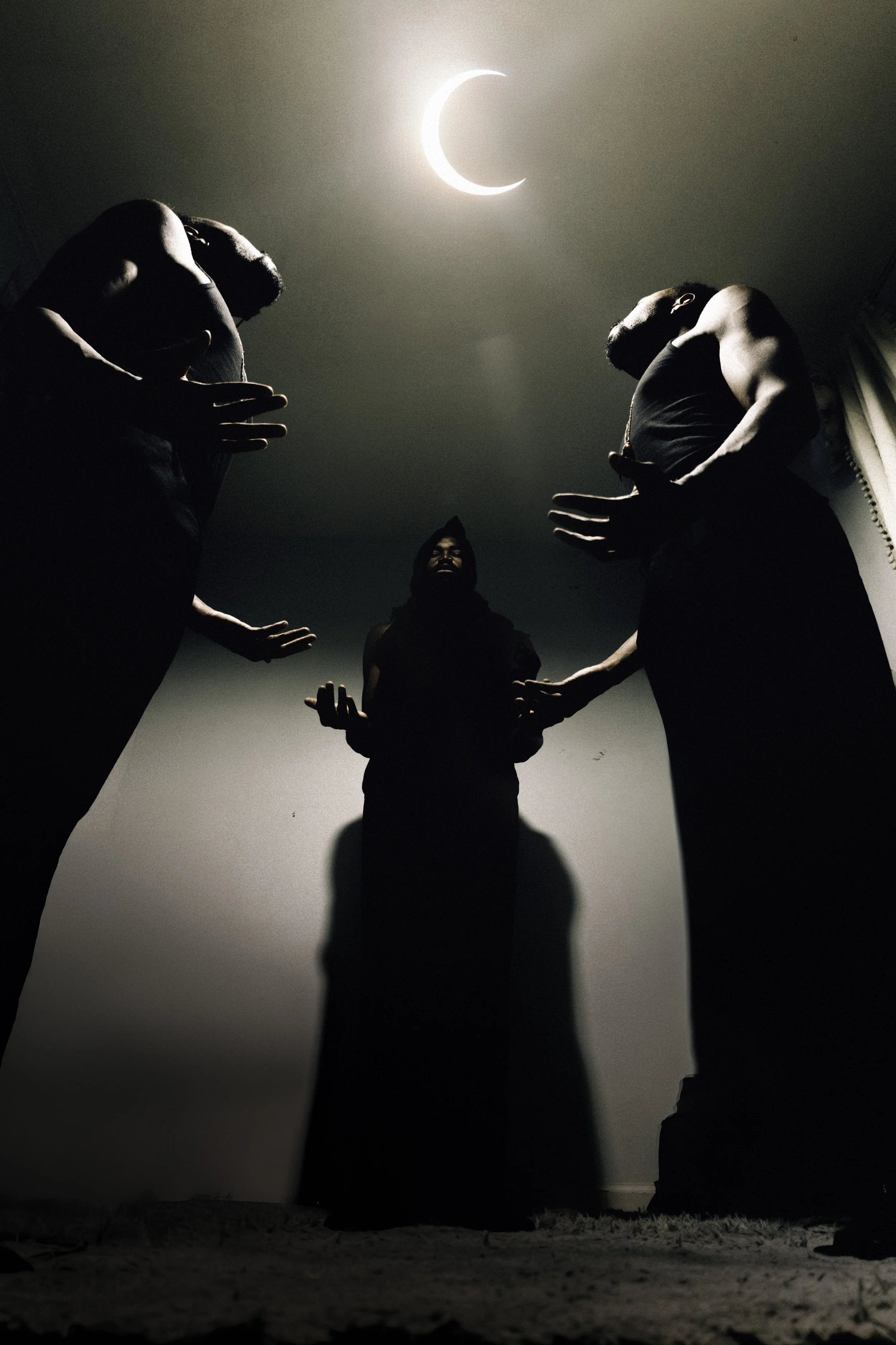 Three people standing in a dark room under a crescent moon-shaped light, with their faces not visible, one person is in the middle facing forward, and the other two are on either side facing inward, all with raised or outstretched hands.
