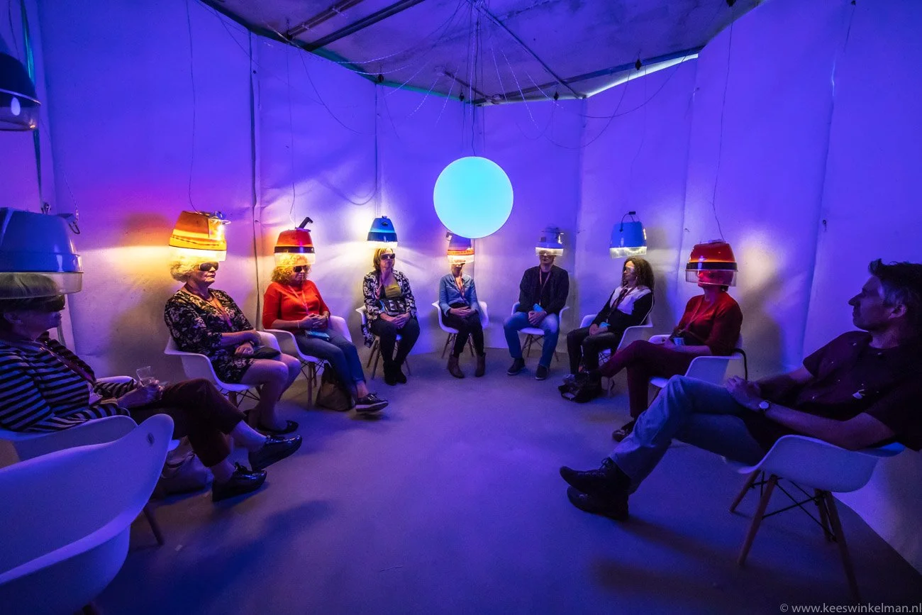 Geledeelnemers zitten in een cirkel in een kamertje met blauwe en paarse verlichting, met een grote blauwe bol in het midden van het plafond en koplampen op hun hoofden die op de muur schijnen.