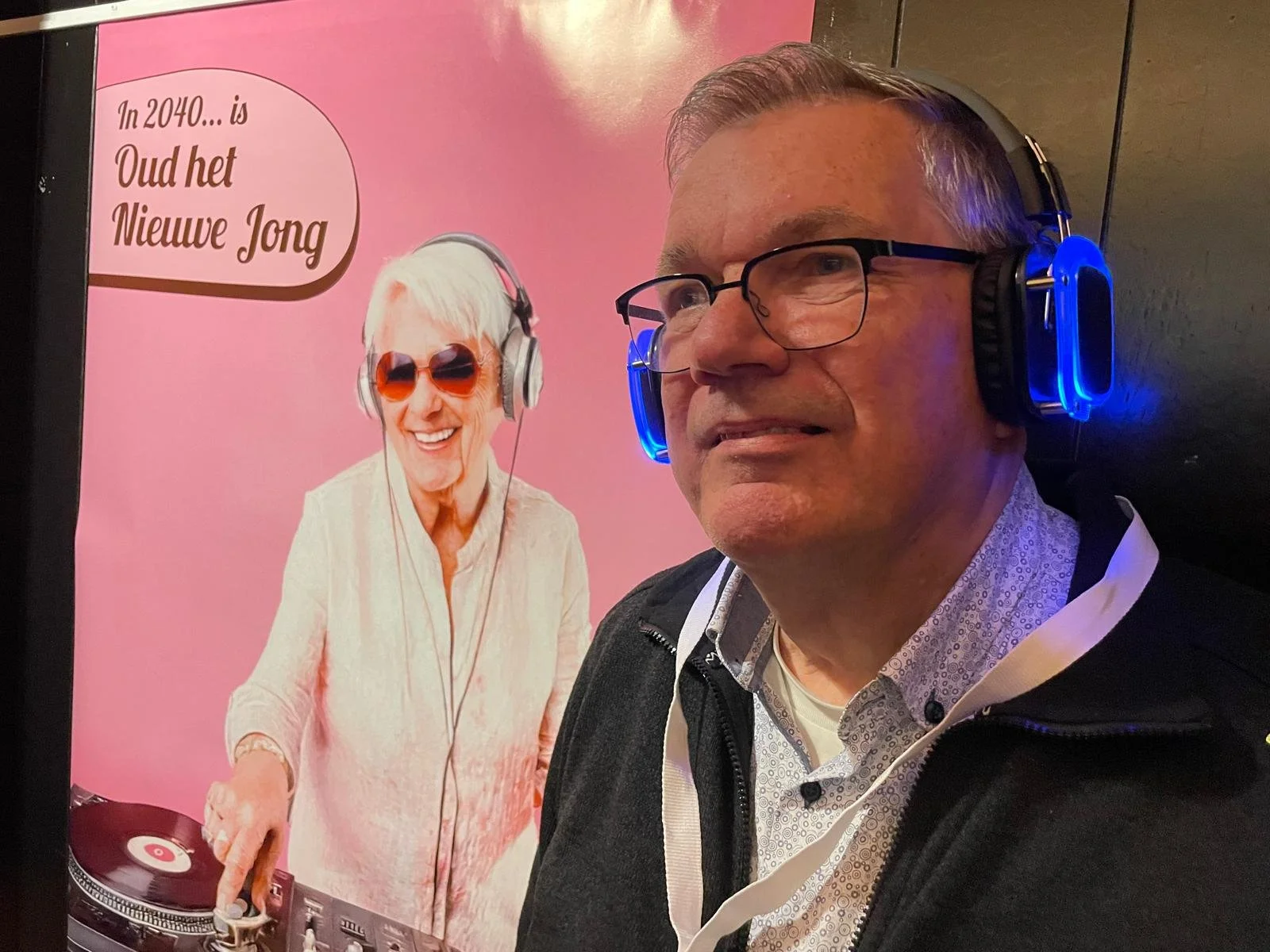 man met bril en koptelefoon voor een poster met een oudere vrouw met koptelefoon en een draaitafel, tekst in het Nederlands, 'In 2040... is Oud het Nieuwe Jong'.