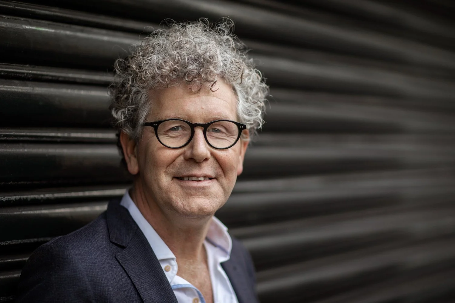 Portret van een volwassen man met grijs, gekruld haar en zwarte bril, lachend, voor een zwarte metallische achtergrond.