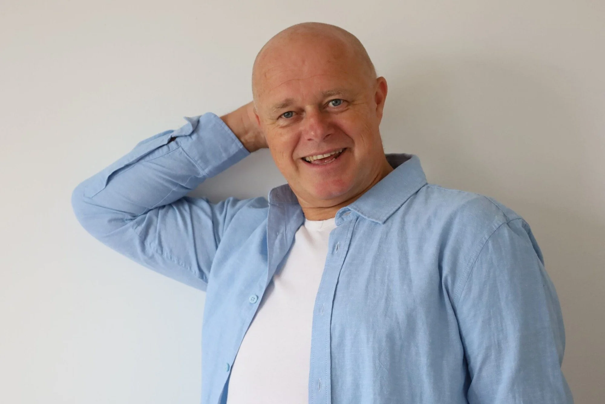 Een man met een blauwe overhemd en wit t-shirt lacht, met zijn rechterhand achter zijn hoofd, voor een witte achtergrond.