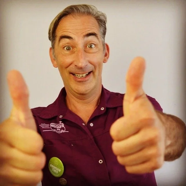 Man met grijs haar in een paarse polo geeft tweemaal duim omhoog en glimlacht naar de camera.