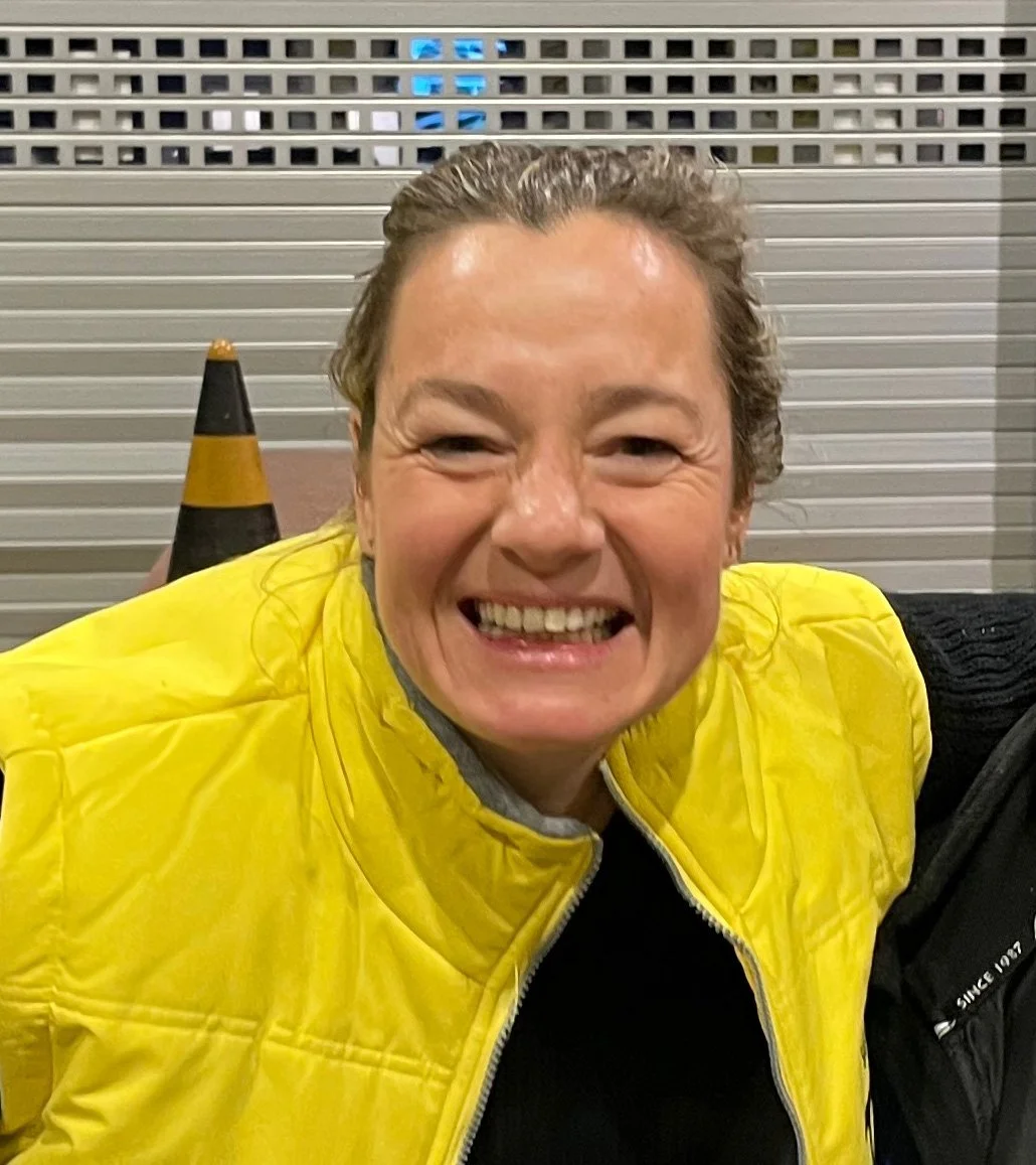 Een lachende vrouw met een geel jasje naast een verkeerskegel en een metalen muurachtergrond.