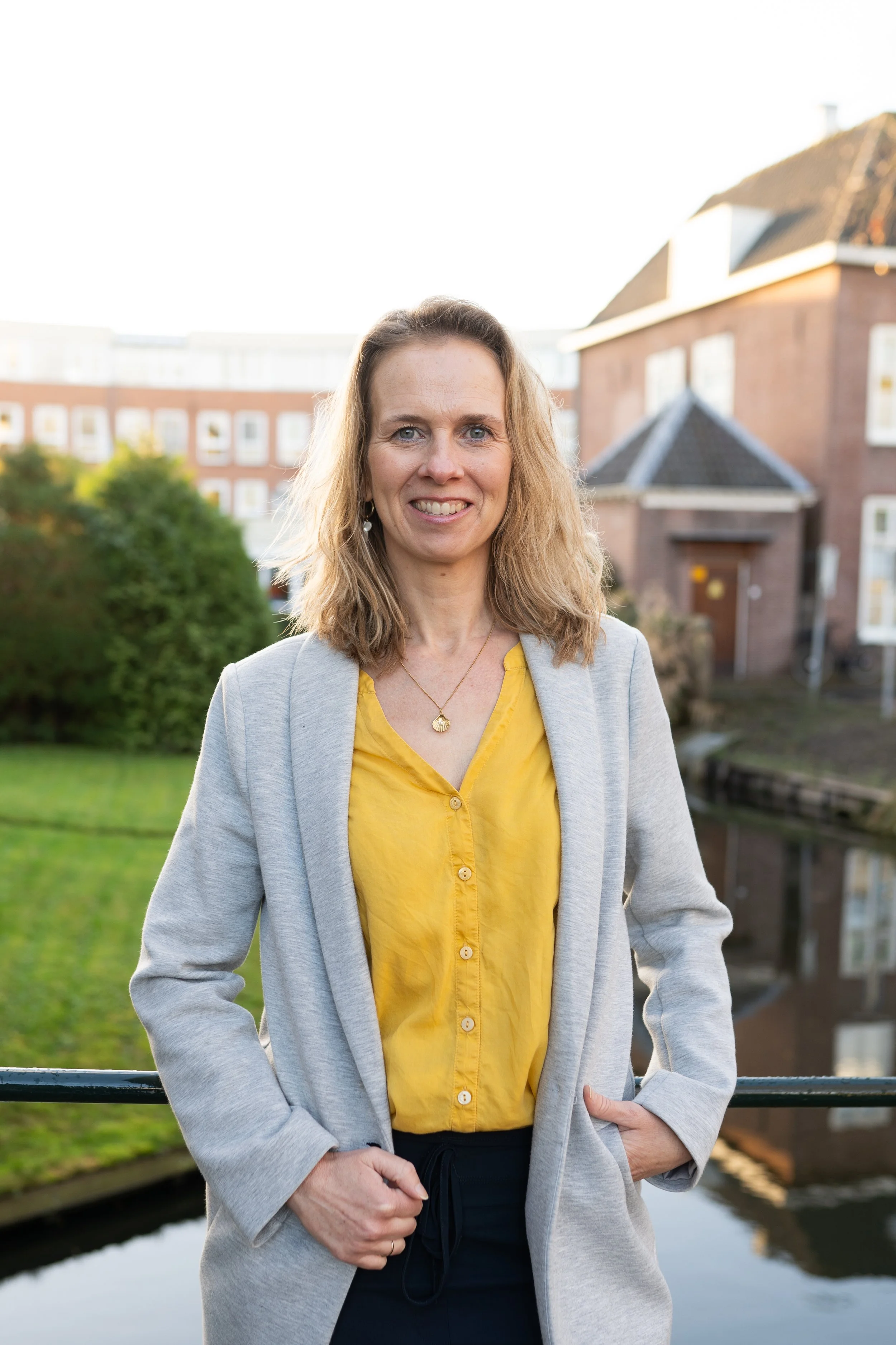 Vrouw in een grijze jas met gele blouse, lachend, staan voor een kanaal met huizen op de achtergrond