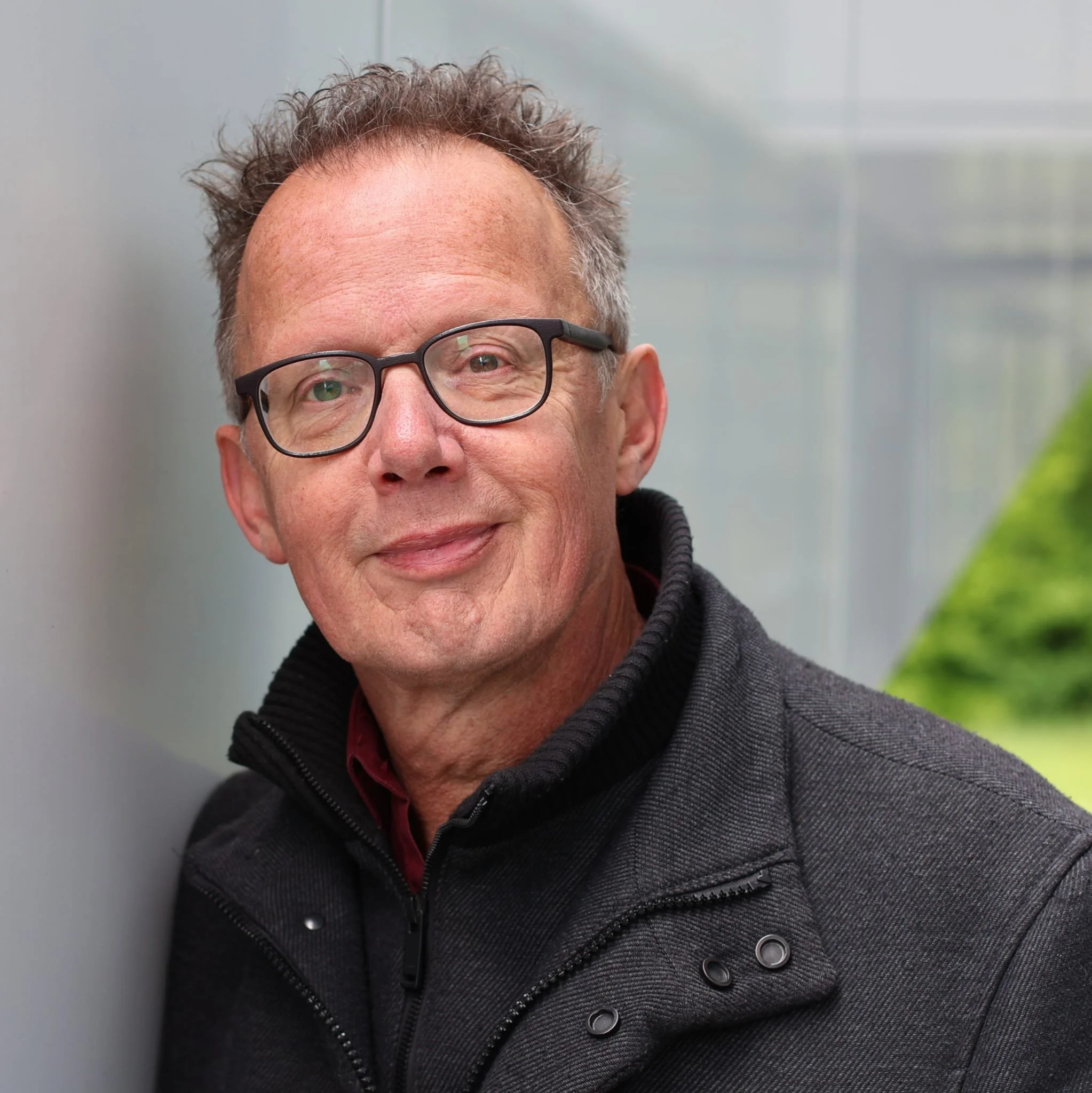 Portret van een glimlachende oudere man met bril in een zwarte jas, staand bij een raam met een groene tuin op de achtergrond.