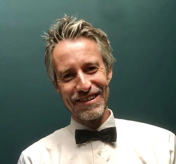 Man met grijze haar en baard, lachend, gekleed in een wit overhemd en een zwarte vlinderstrik, voor een groene achtergrond.