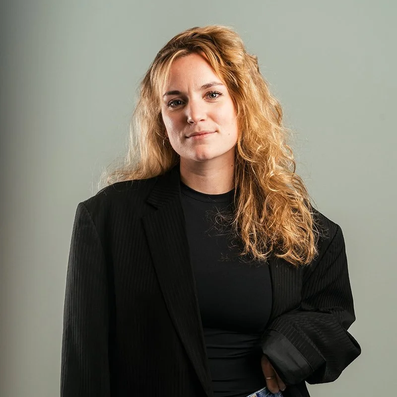 Een jonge vrouw met lang, golvend, rood haar draagt een zwarte blazer en staat voor een effen groene achtergrond.