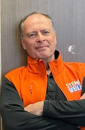 Man met korte grijze haar en een serieuze uitdrukking, dragende een oranje vest met tekst 'Stek Veilig', staand voor een grijze muur.