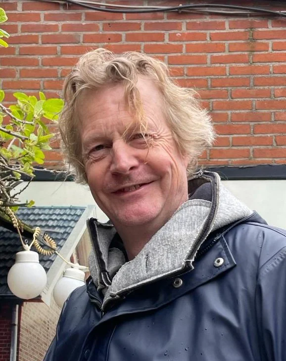 Een lachende man met warrig blond haar, gekleed in een grijze hoodie en een blauwe regenjas, staat buiten voor een bakstenen muur.