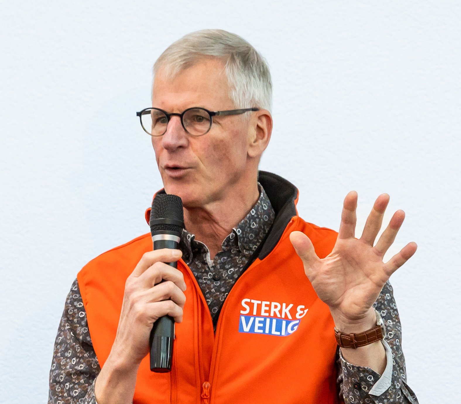 Man met grijs haar en bril, die een microfoon vasthoudt en spreekt, draagt een oranje vest met de tekst 'Sterk & Veilig'.