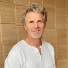 Portret van een man met grijs haar en een lichte baard, gekleed in een witte shirt, staand voor een houten achtergrond.