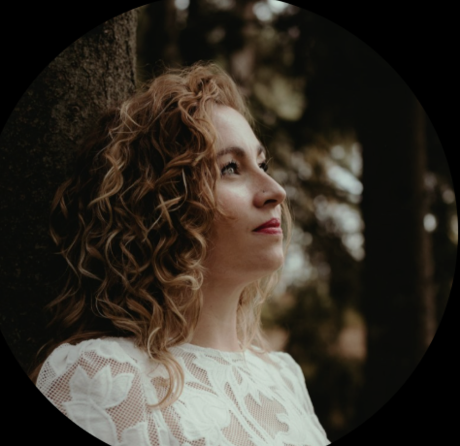 Portret van een vrouw met krullend, lichtbruin haar, rode lippen, in een witte lace top, staand buiten bij bomen.