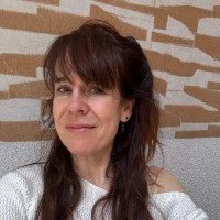 Portret van een vrouw met bruin haar en een witte trui, zittend voor een bakstenen muur.