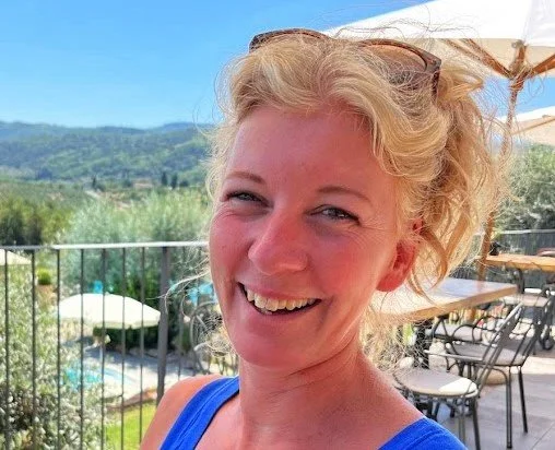 Een lachende vrouw met blond haar op een terras met uitzicht op een tuin en bergen, onder een wit parasol.