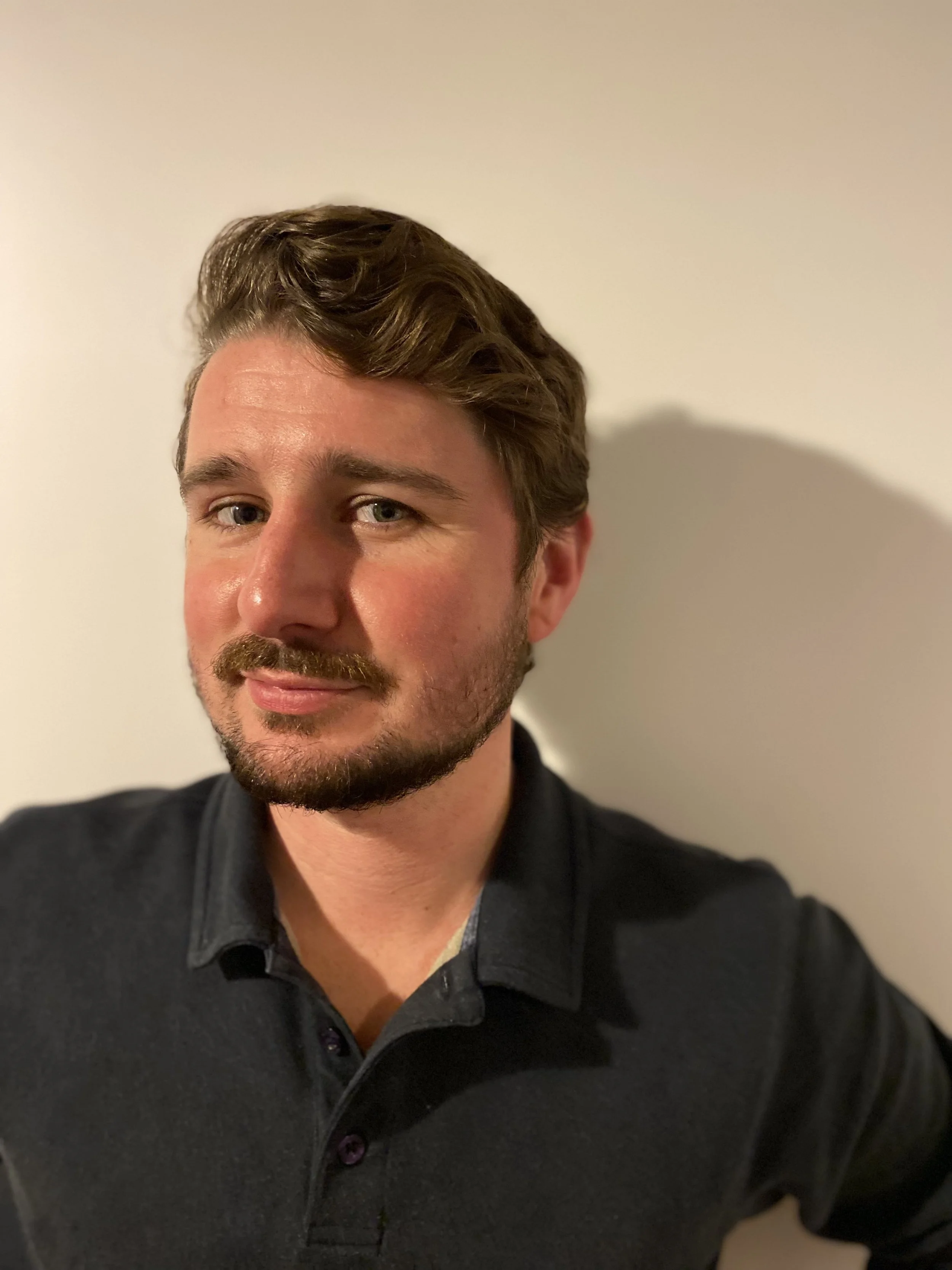 Portret van een man met bruin golvend haar, een vollere baard en een lichte huid, die glimlacht en gekleed is in een donkerkleurige polo, staand tegen een effen lichte achtergrond.