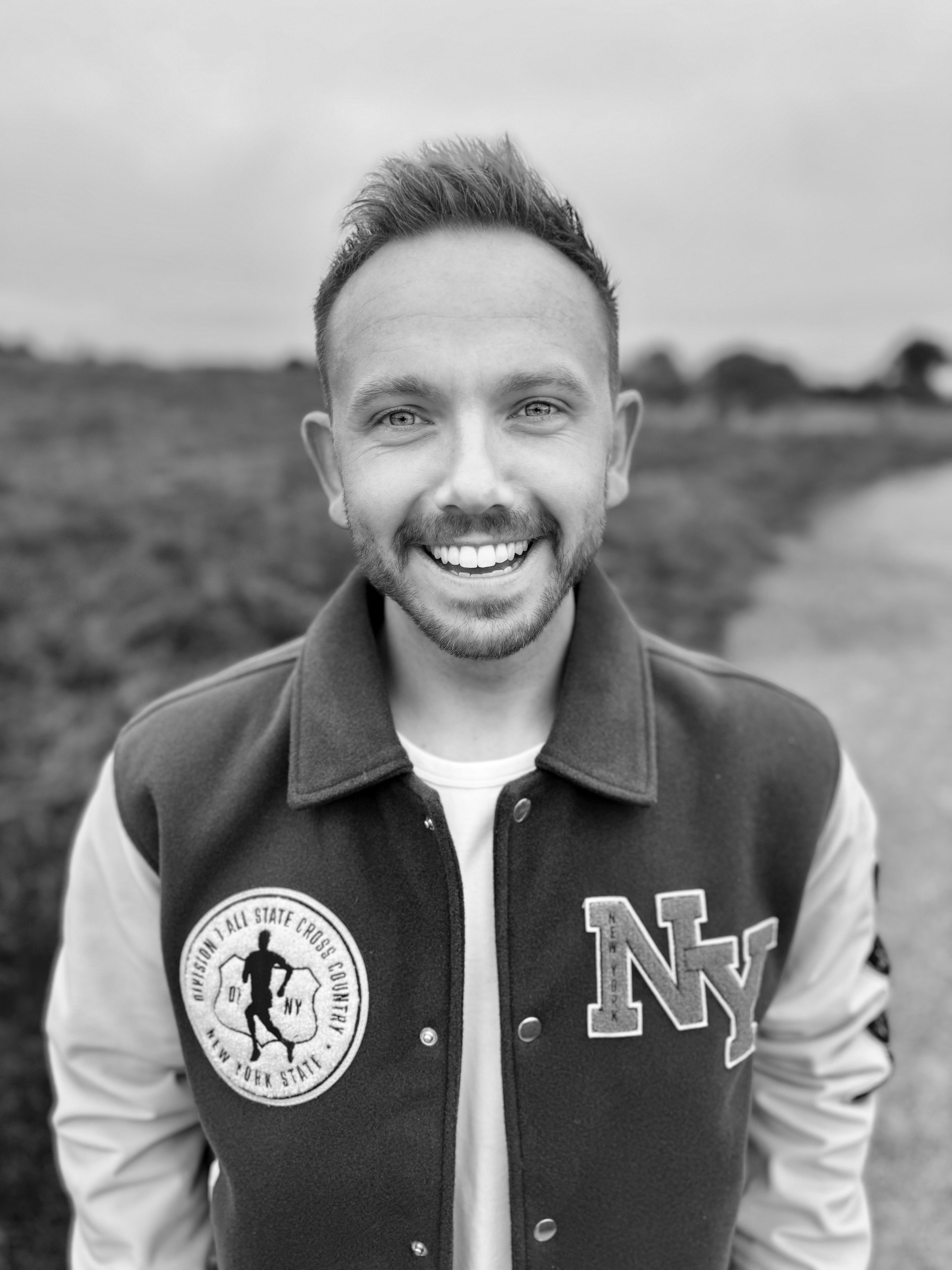 A smiling young man with short hair and a beard, wearing a varsity jacket with patches, standing outdoors in a natural setting with trees and a cloudy sky in the background.