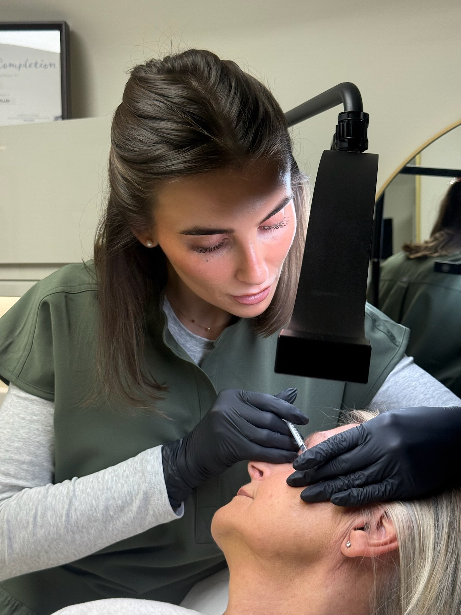 A woman with long brown hair receiving a tattoo on her hand from a tattoo artist wearing gloves, in a studio setting.