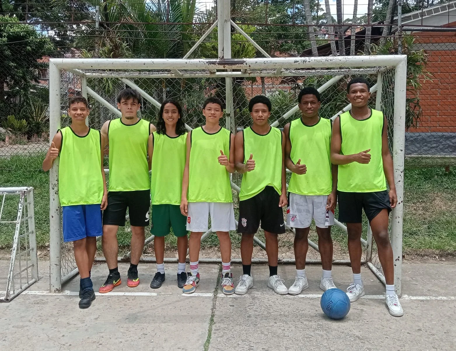 Grupo de siete jóvenes deportistas en uniforme de entrenamiento, de pie frente a una portería de fútbol, todos sonriendo y haciendo gestos positivos, en un campo de concreto con área verde y una pelota azul al frente.