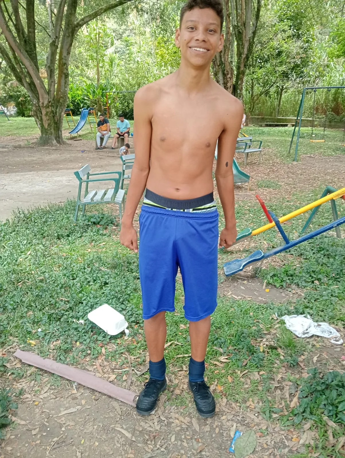 Un joven con pantalones cortos azules y sin camiseta en un parque con columpios y toboganes, sonriendo a la cámara.