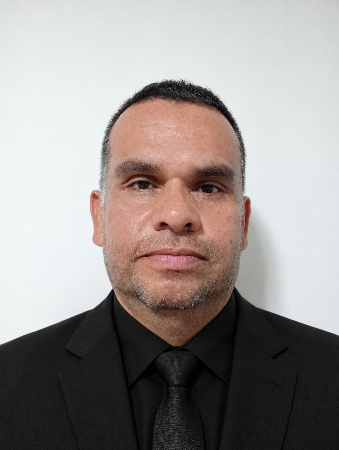 Retrato de un hombre con barba, rasgos oscuros y cabello corto, vestido con traje negro y corbata negra, en fondo blanco.