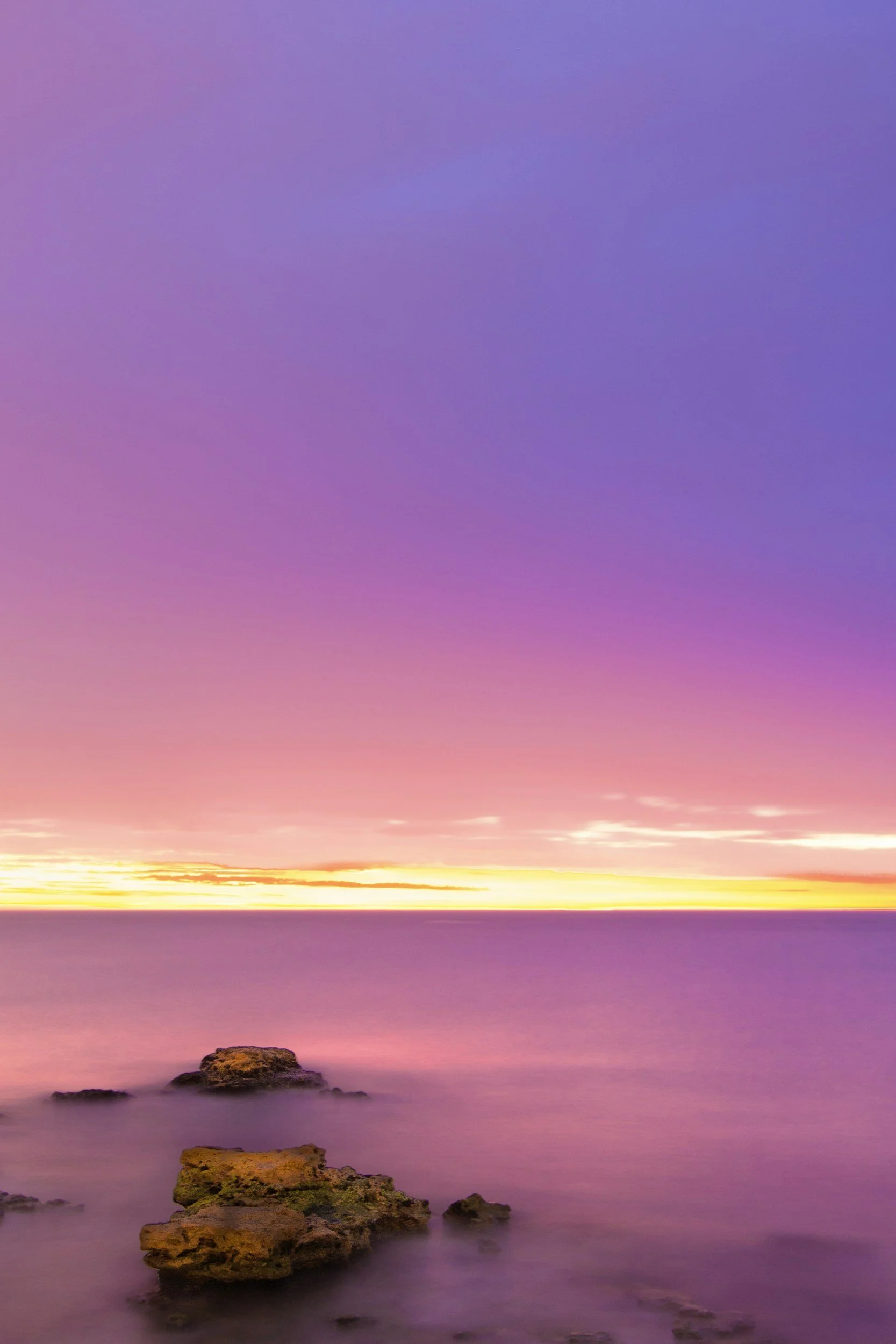 Colorful sunset over calm water with rocks in the foreground and a vibrant gradient sky.