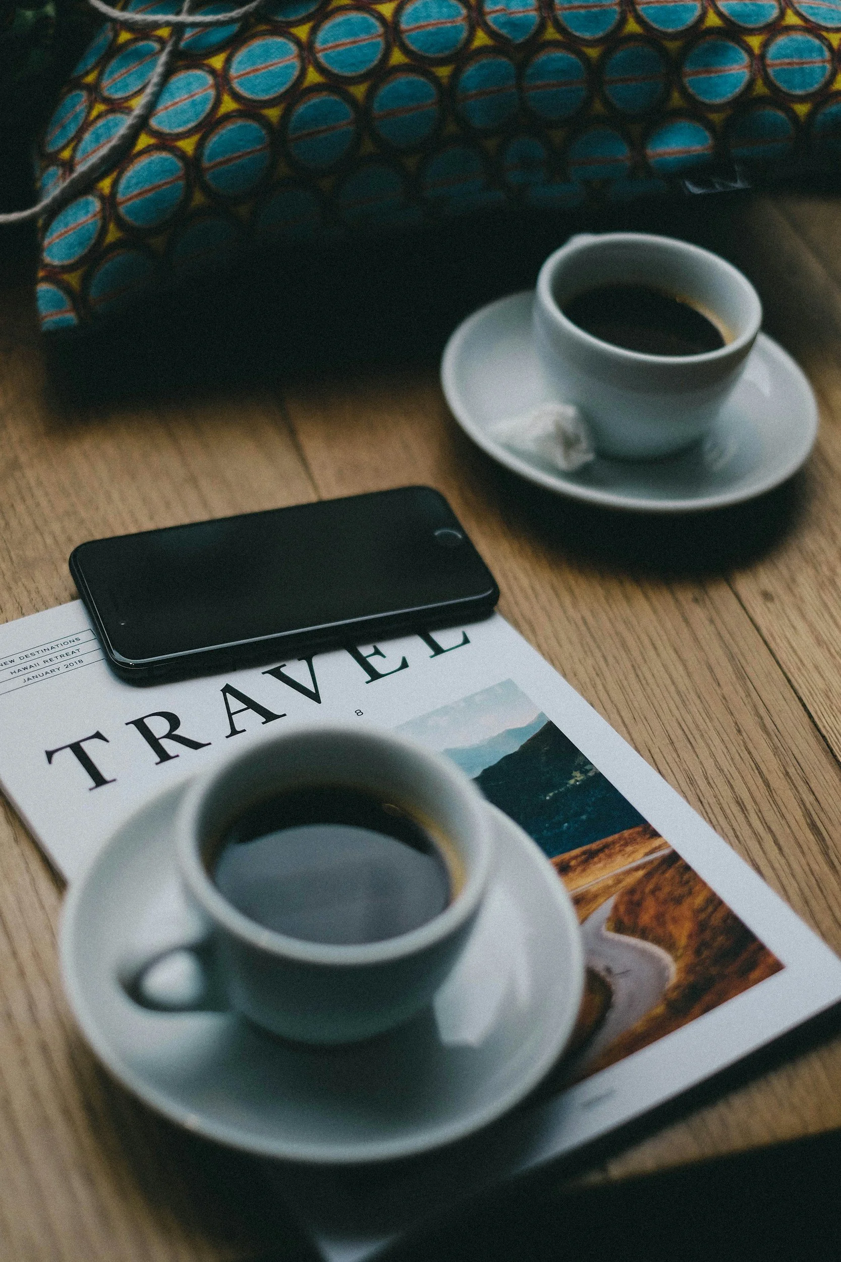 A cup of black coffee on a saucer with a silver spoon, placed on a wooden table next to a travel magazine and a smartphone, with a colorful patterned pouch in the background.