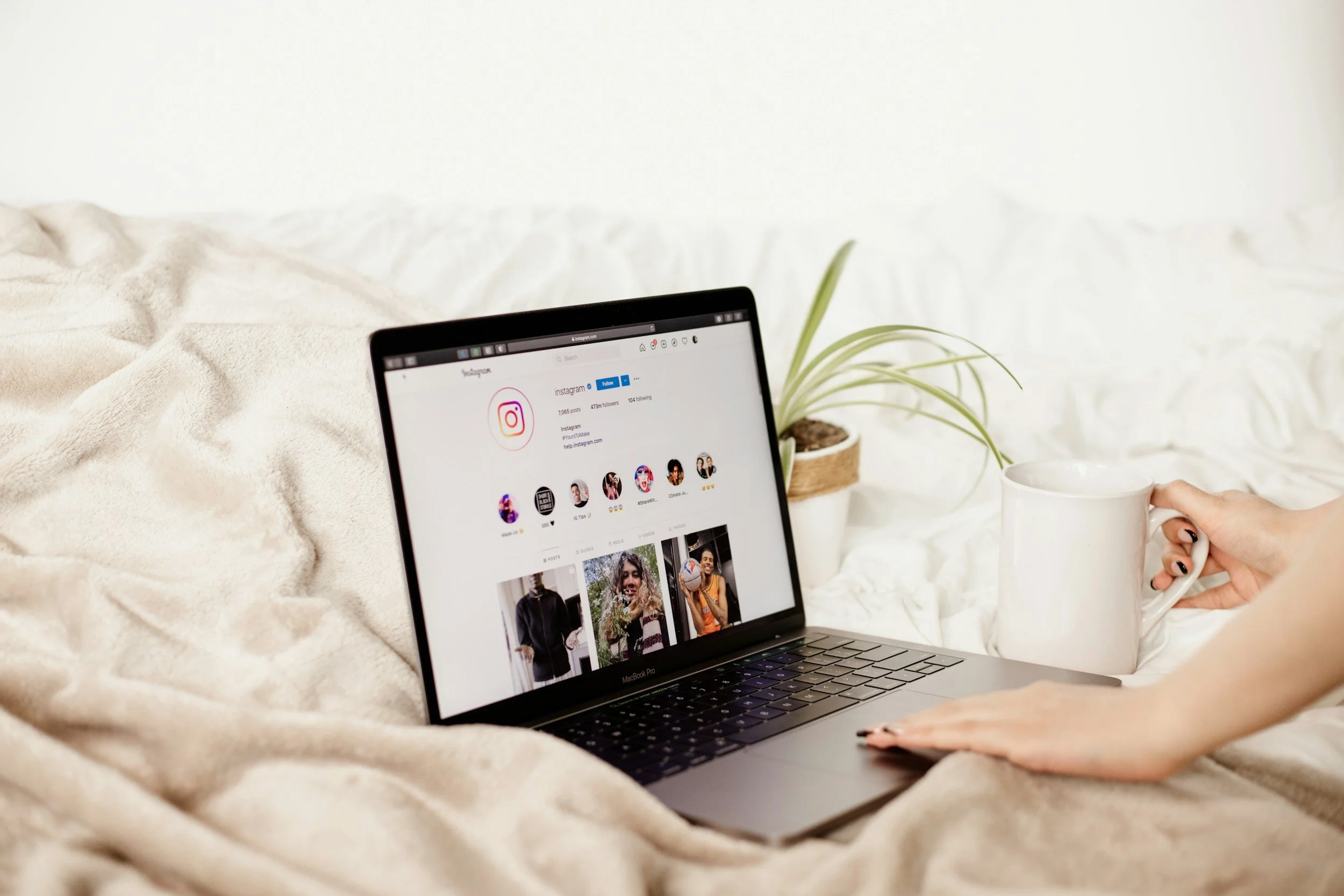A person holding a white mug, sitting on a bed with a beige blanket, with a MacBook Pro laptop on the bed displaying an Instagram profile page.