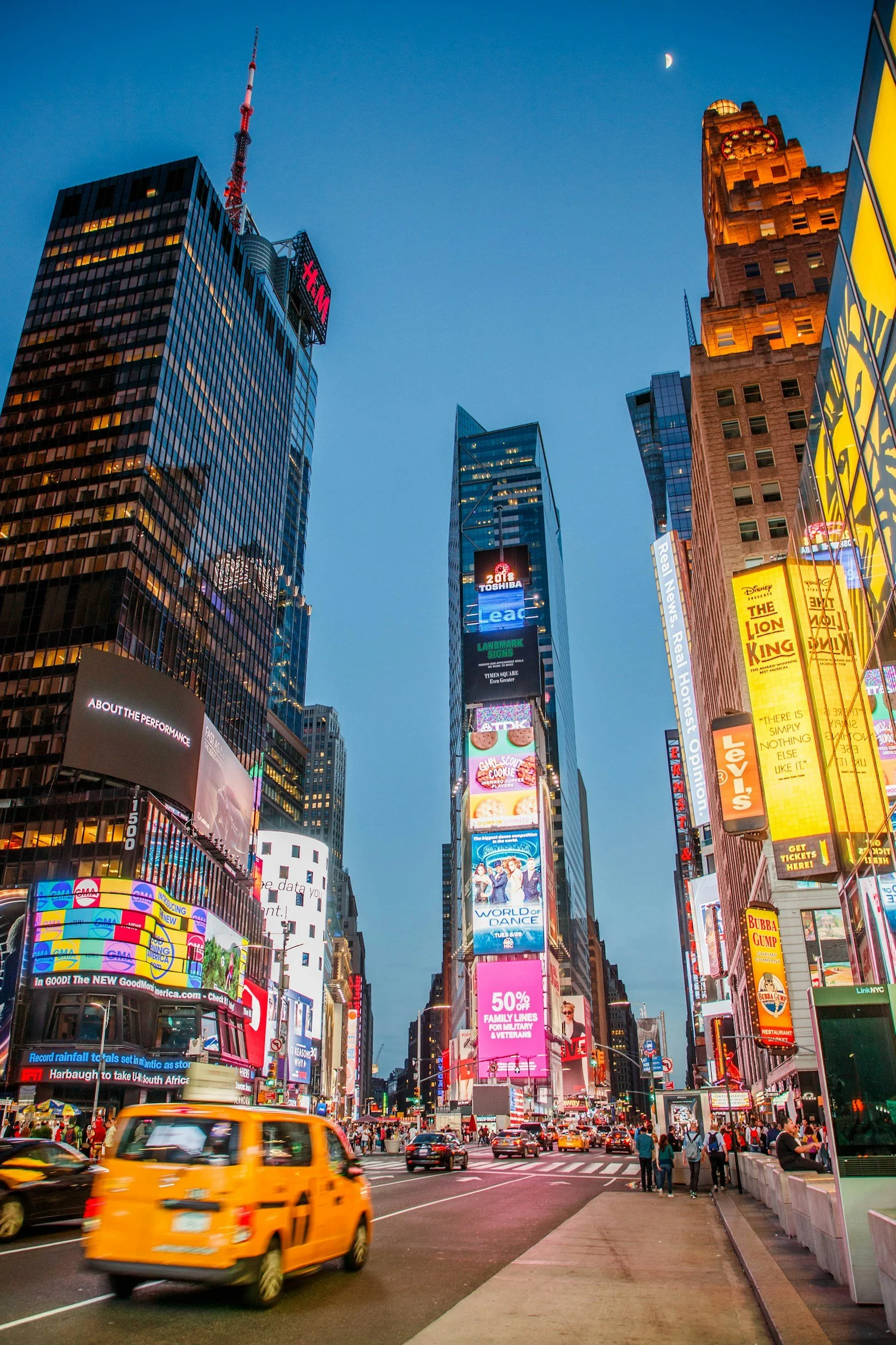 Nighttime view of Times Square in New York City with tall illuminated billboards, traffic including a yellow taxi, and pedestrians on the sidewalk.