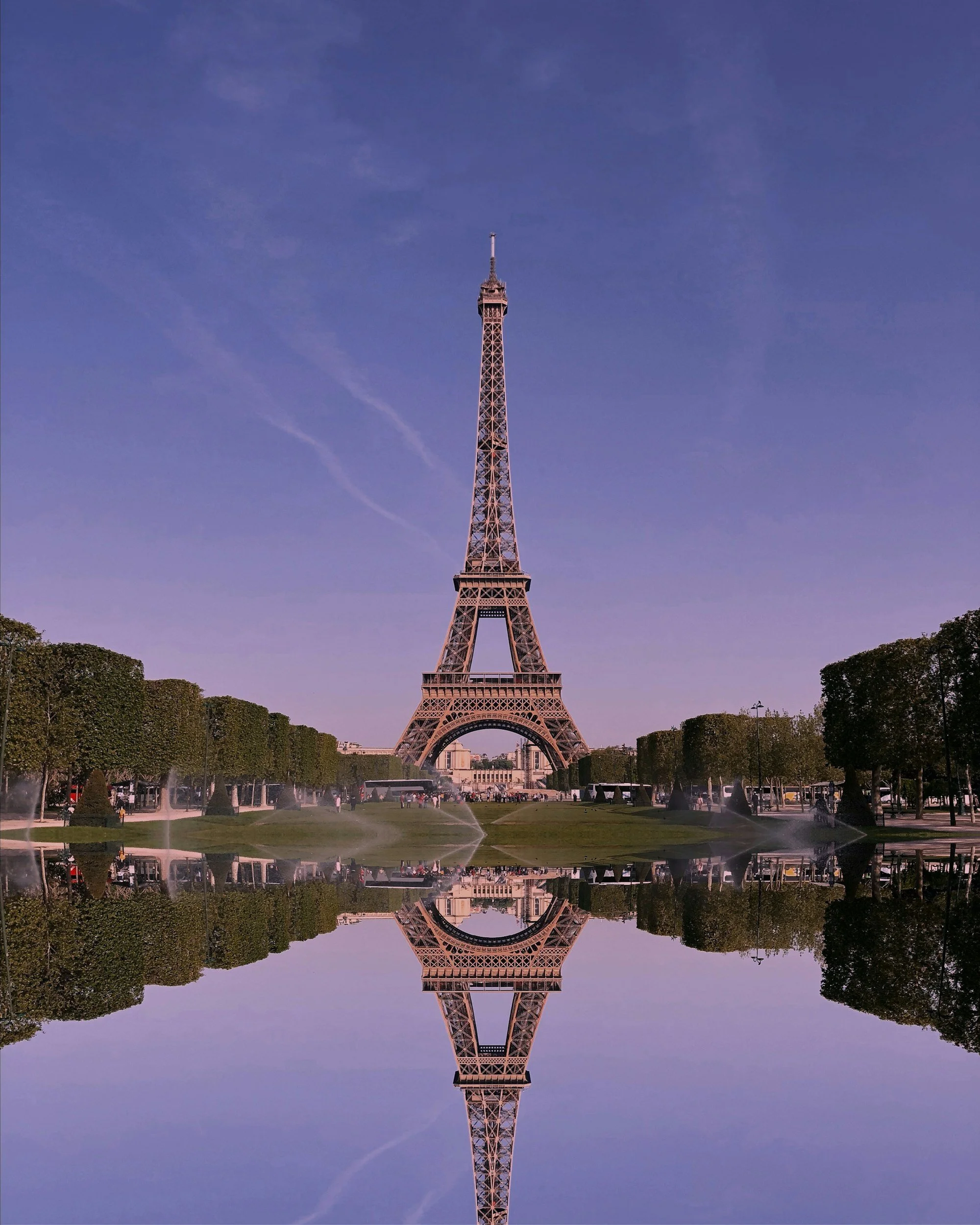 The Eiffel Tower in Paris, France, with a reflection in a body of water at its base, greenery around, and a partly cloudy sky.
