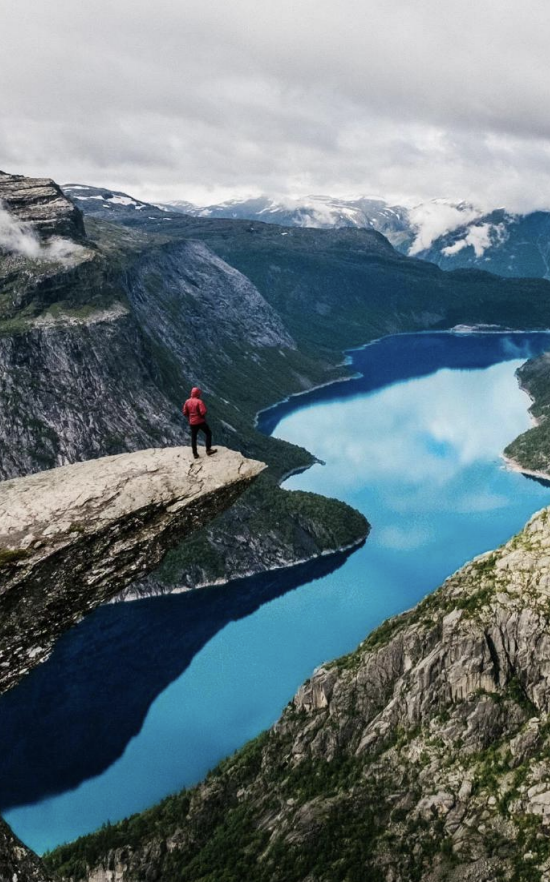 A person in a red jacket standing on a large rock protruding from a cliff overlooking a blue lake surrounded by mountains.