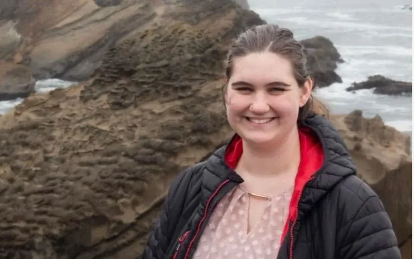 A young woman with long brown hair tied back, smiling, wearing a pink polka-dot blouse and a black jacket with a red lining, standing on rocky coastal terrain with the ocean in the background.