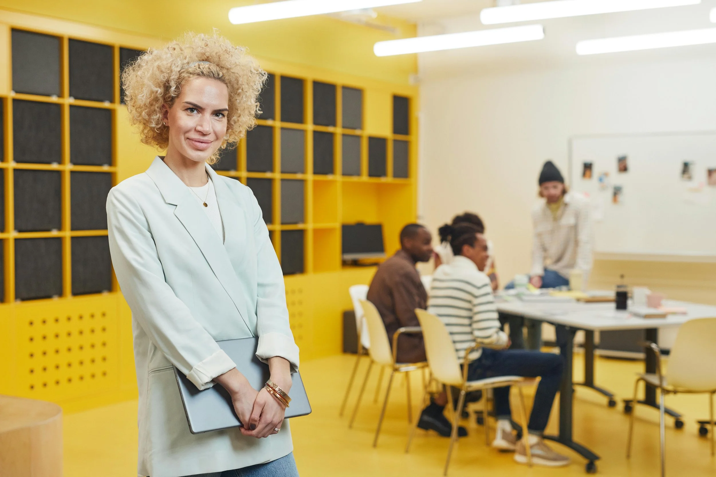 A woman with curly blonde hair and light skin standing in a classroom with yellow walls and black wall squares, holding a laptop. In the background, students are sitting at tables and a person with a beanie stands smiling.