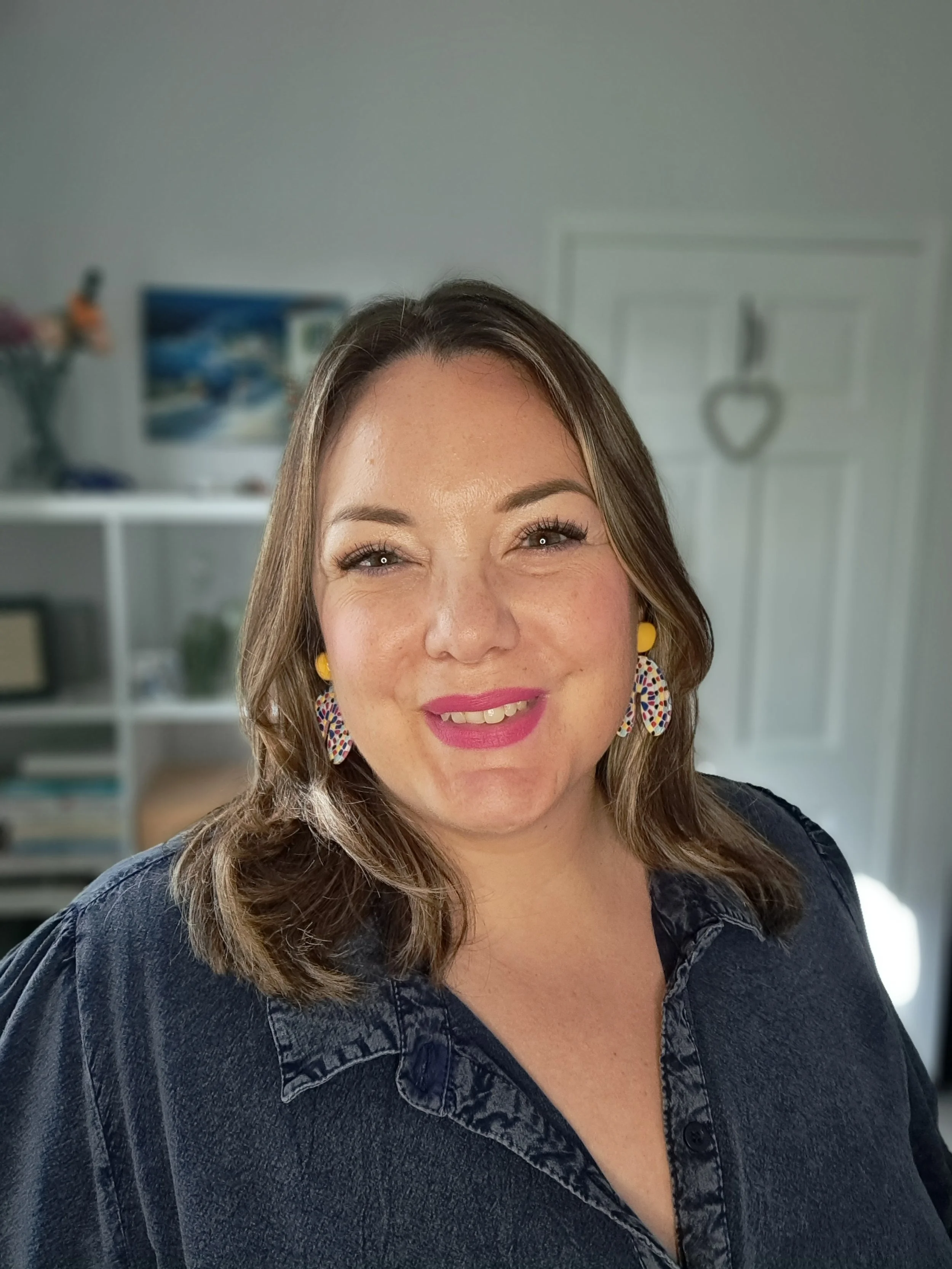 A smiling woman with brown hair wearing yellow earrings and a dark denim shirt, indoors with a blurred background of shelves and a heart-shaped decoration on the door.