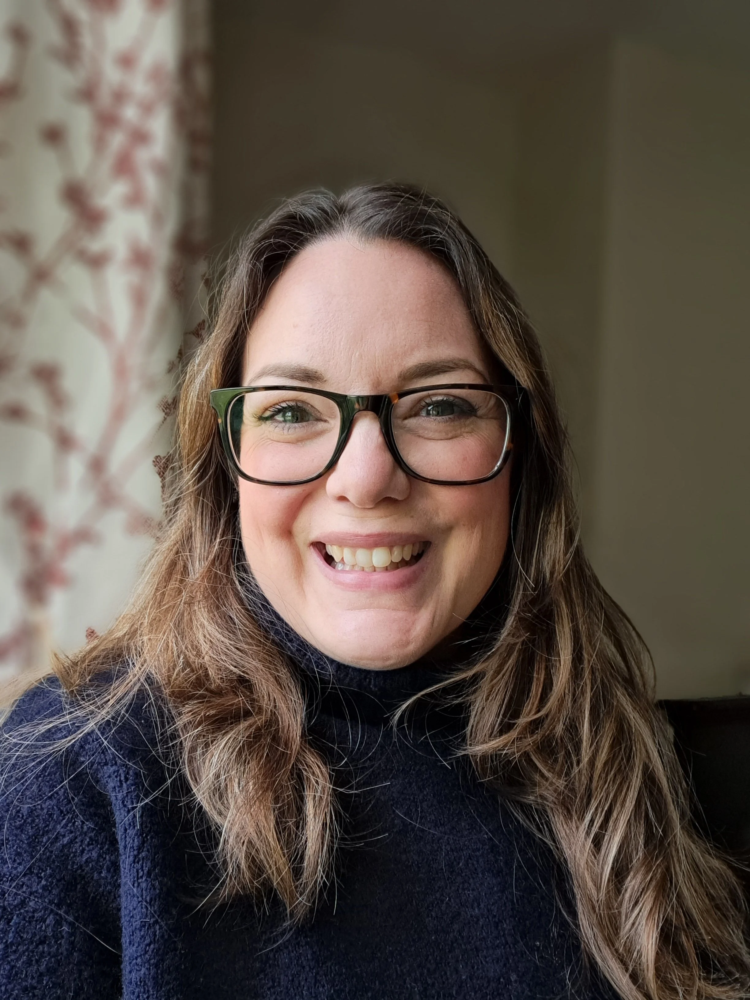 Close-up of a smiling woman with long, wavy hair, wearing glasses, in a dimly lit room with patterned curtains.