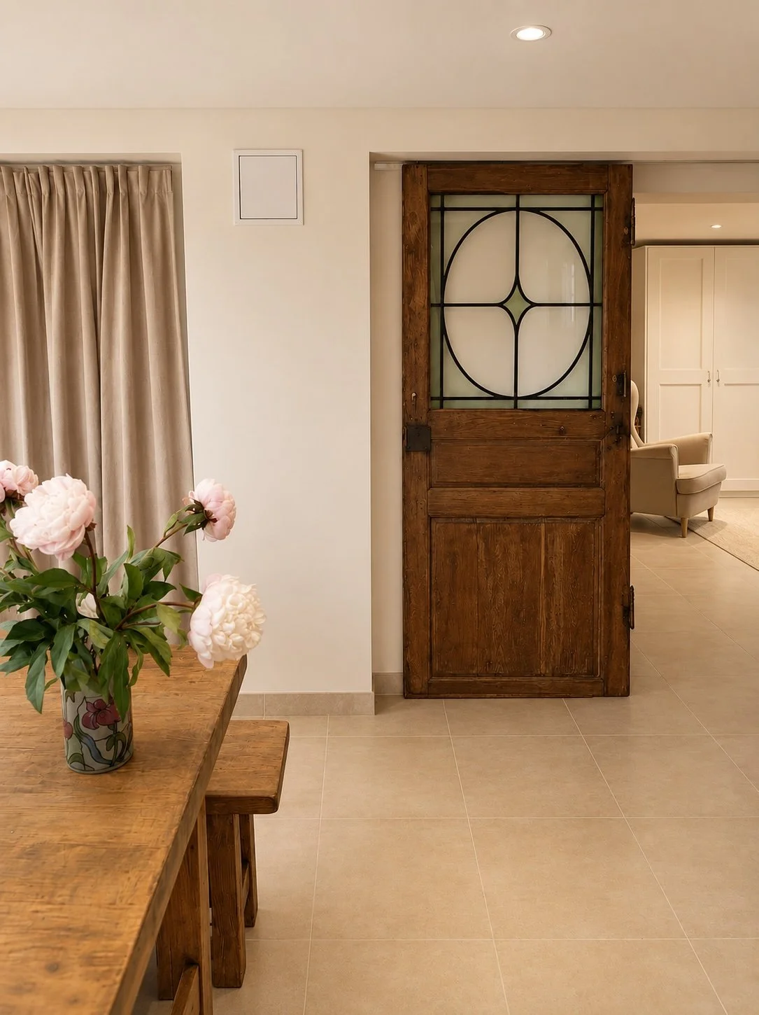 Une pièce intérieure avec un mur blanc, une porte en bois avec un panneau en verre orné d'un motif géométrique noir, un tapis beige, une chaise beige, et un meuble blanc en fond. Un vase avec des pivoines roses est posé sur une table en bois près d'un rideau beige.
