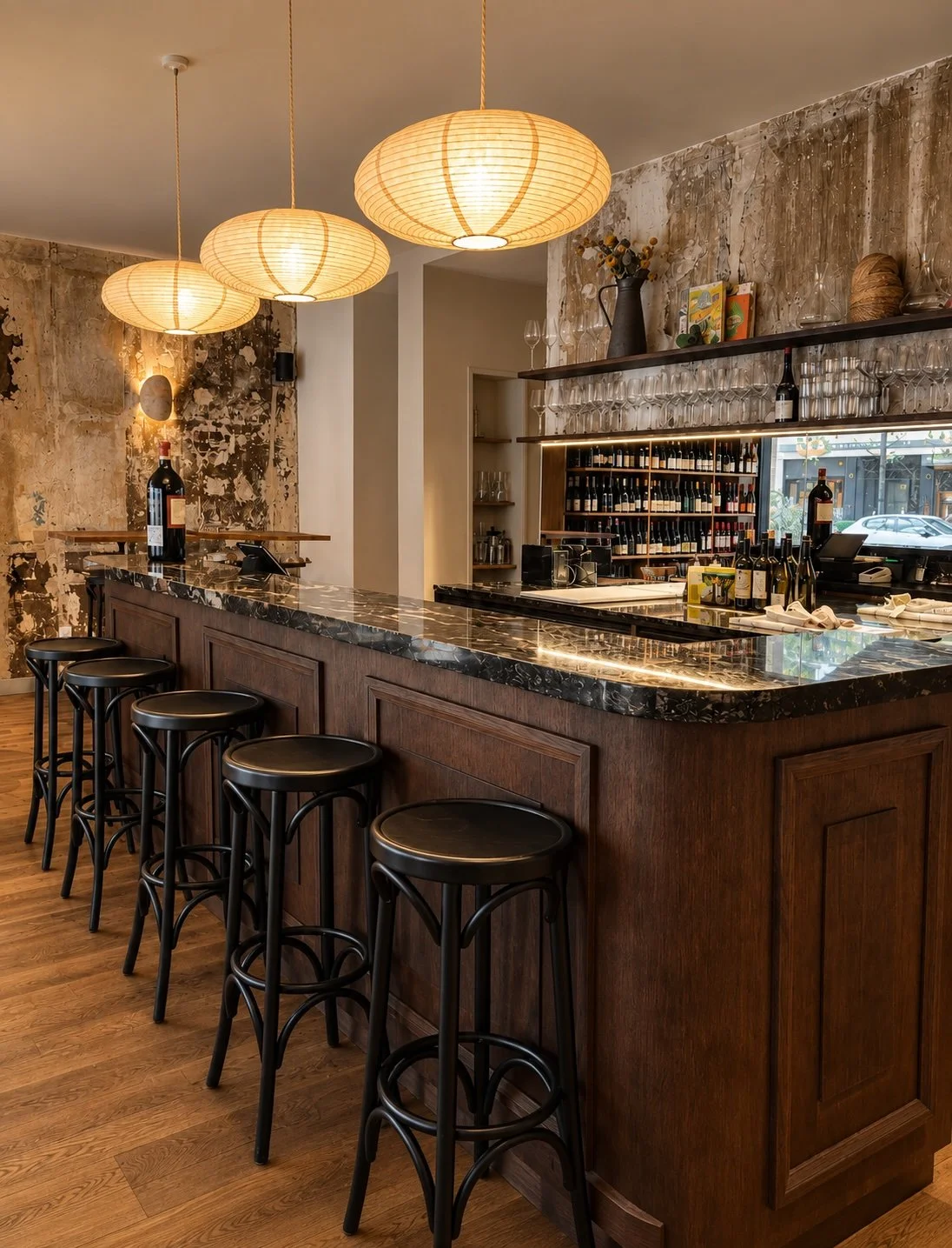 Bar avec comptoir en marbre noir, chaises hautes en bois et métal, étagère avec bouteilles de vin et verres, éclairage suspendu en papier, murs en béton brut et vieux bois.