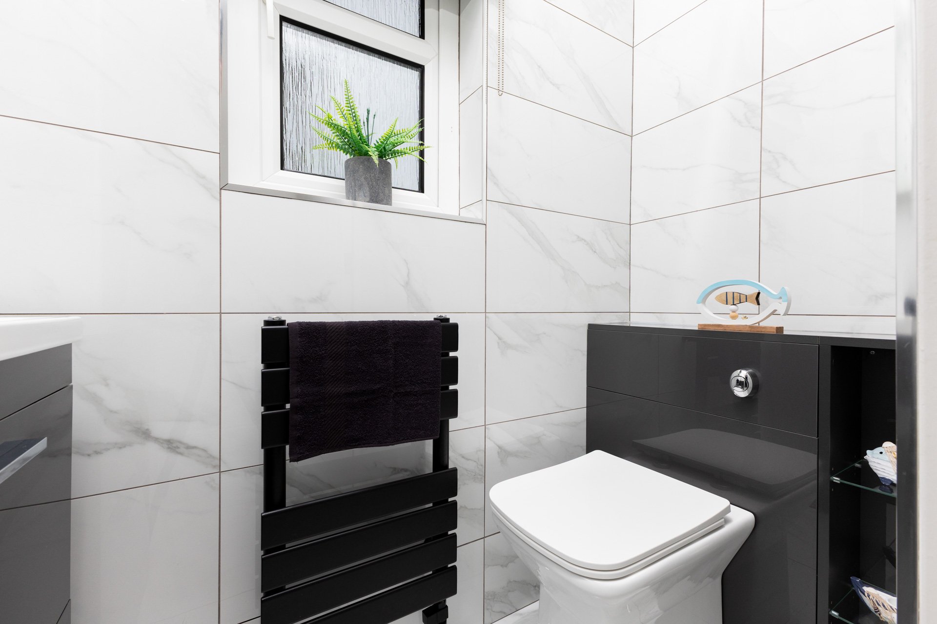 Modern bathroom with white marble tiled walls, a window with frosted glass, a black towel rack with a black towel, and a black cabinet with decorative items, including a fish-shaped ornament.
