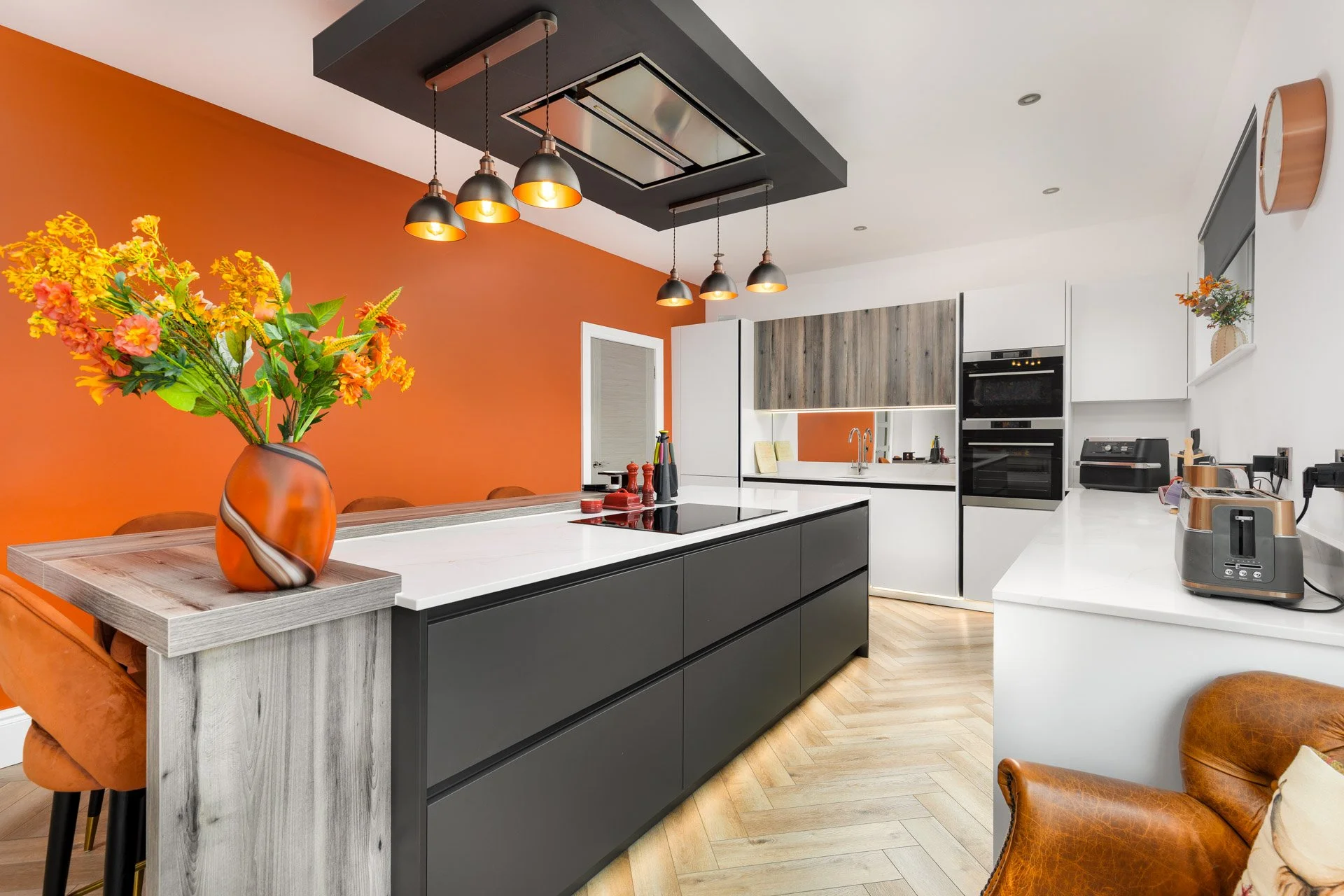 Modern kitchen with orange accent wall, white countertops, black lower cabinets, and light wood upper cabinets. Decor includes a large orange vase with yellow and orange flowers, a leather armchair, and kitchen appliances like a toaster and coffee ma