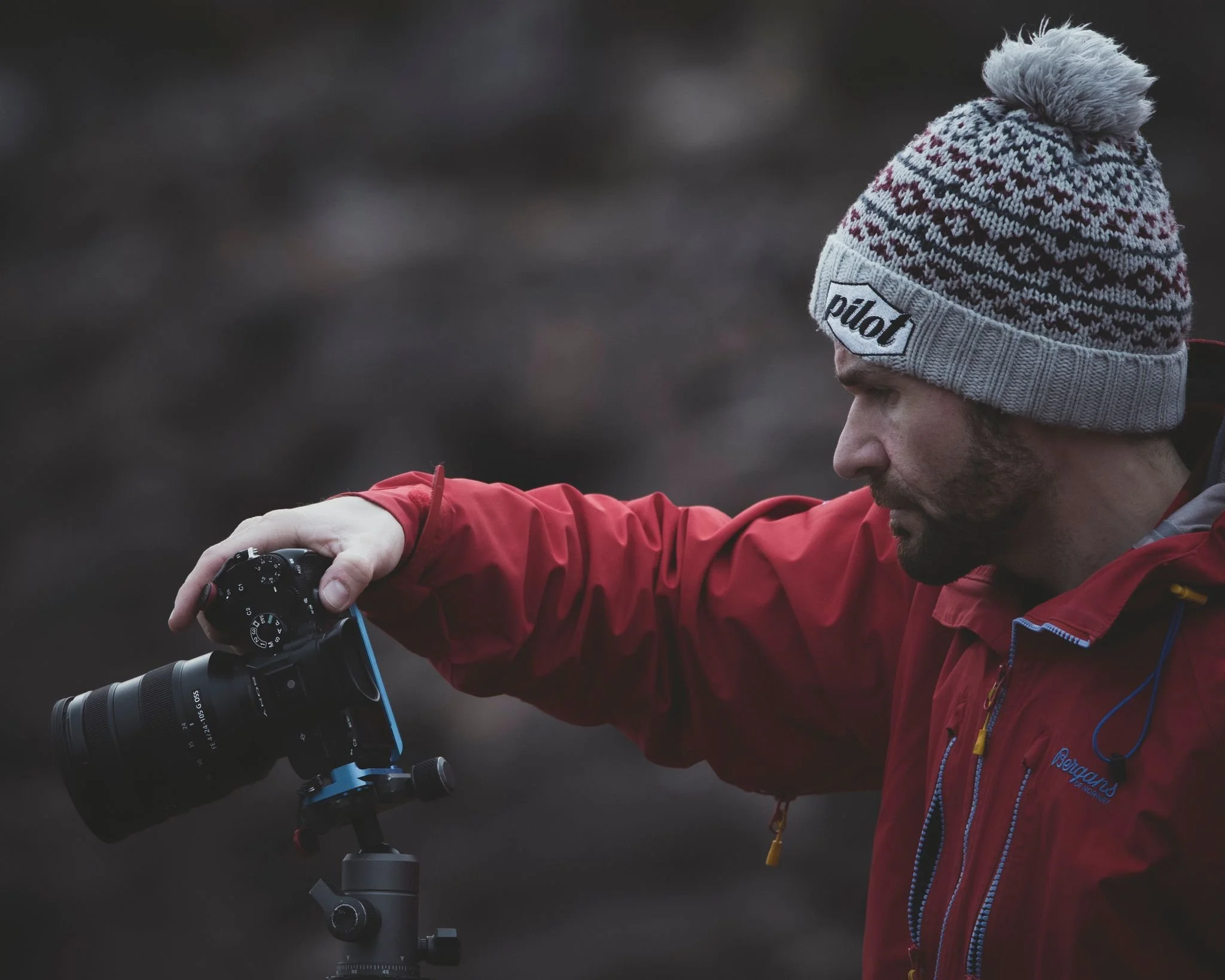 A person wearing a red jacket and a gray, white, and red knitted beanie with a pom-pom on top, adjusting a camera mounted on a tripod outdoors with a blurred background.